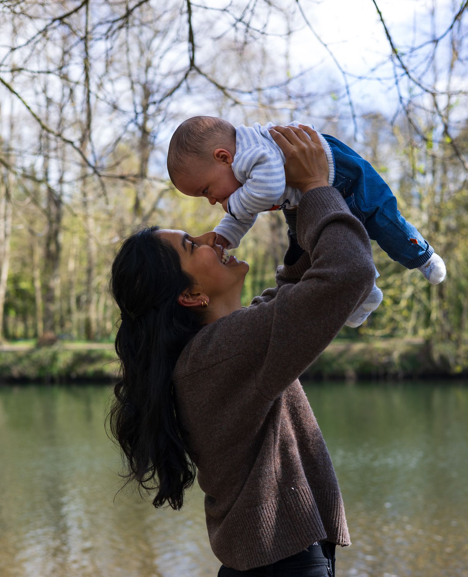 Mother lifting baby up joyfully beside a river during a family photoshoot