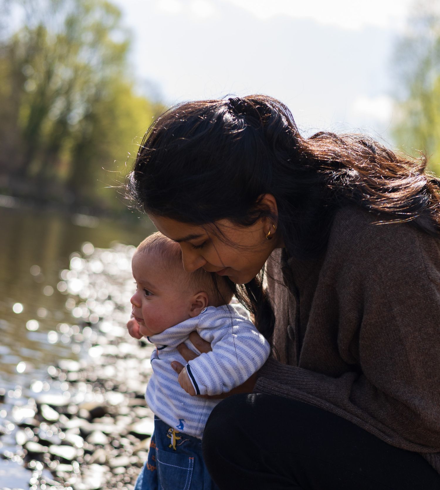 Mother cradling and kissing her baby beside a pebbled riverbank