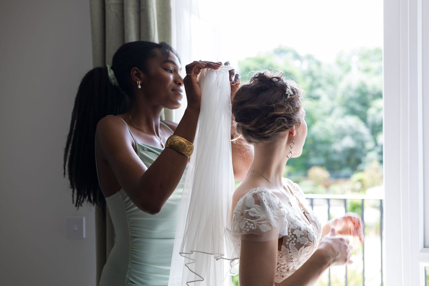 Bridesmaid helping fasten the bride's wedding dress by a window