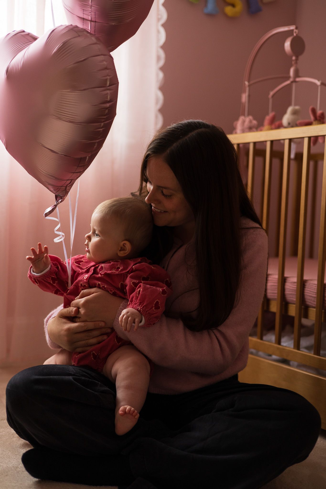 Mother kneeling with her baby beside pink heart balloons at a first birthday party