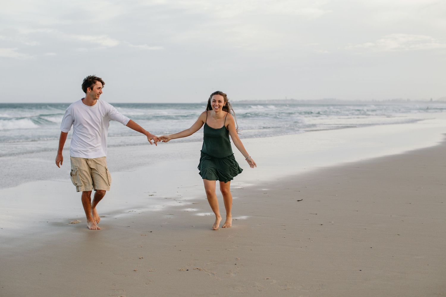 A happy couple holds hands while walking barefoot along a sandy beach with ocean waves in the background.