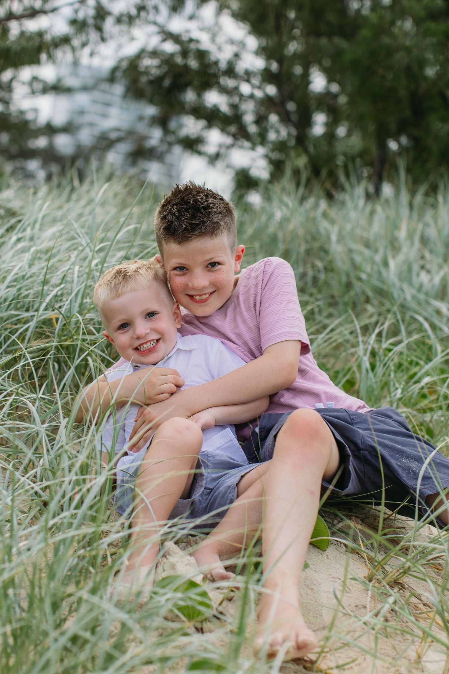 Two brothers smile and hug while sitting together in tall beach grass near the shoreline.