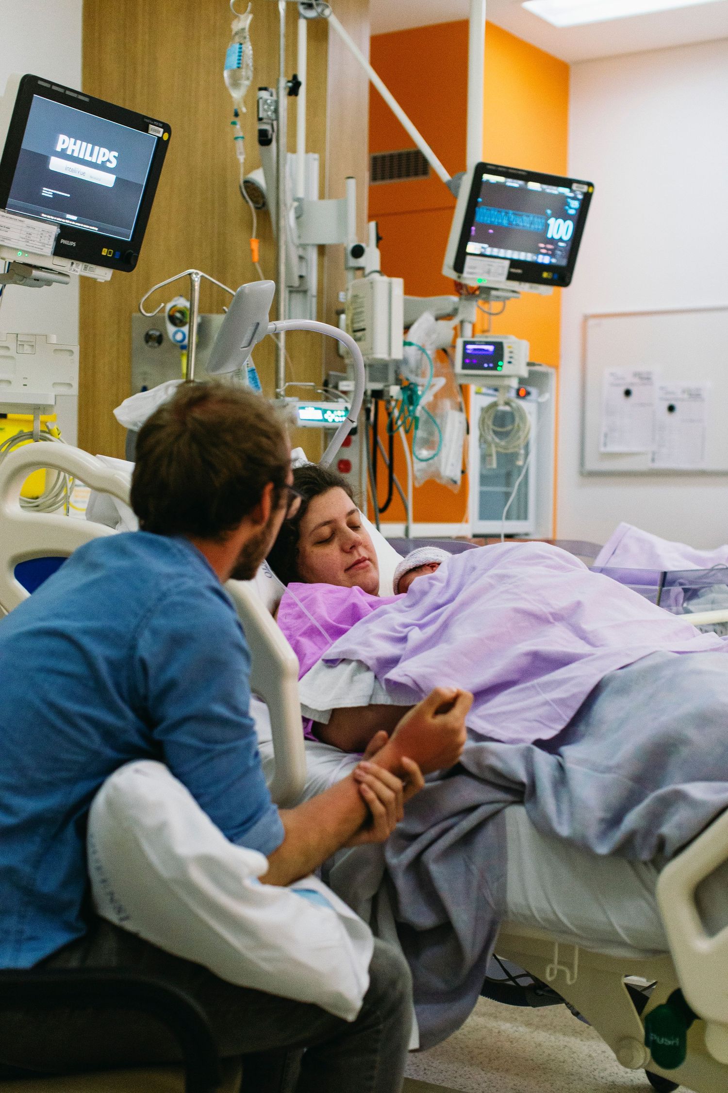 Man sitting beside woman in hospital bed, holding hands surrounded by medical monitors in a modern hospital room.