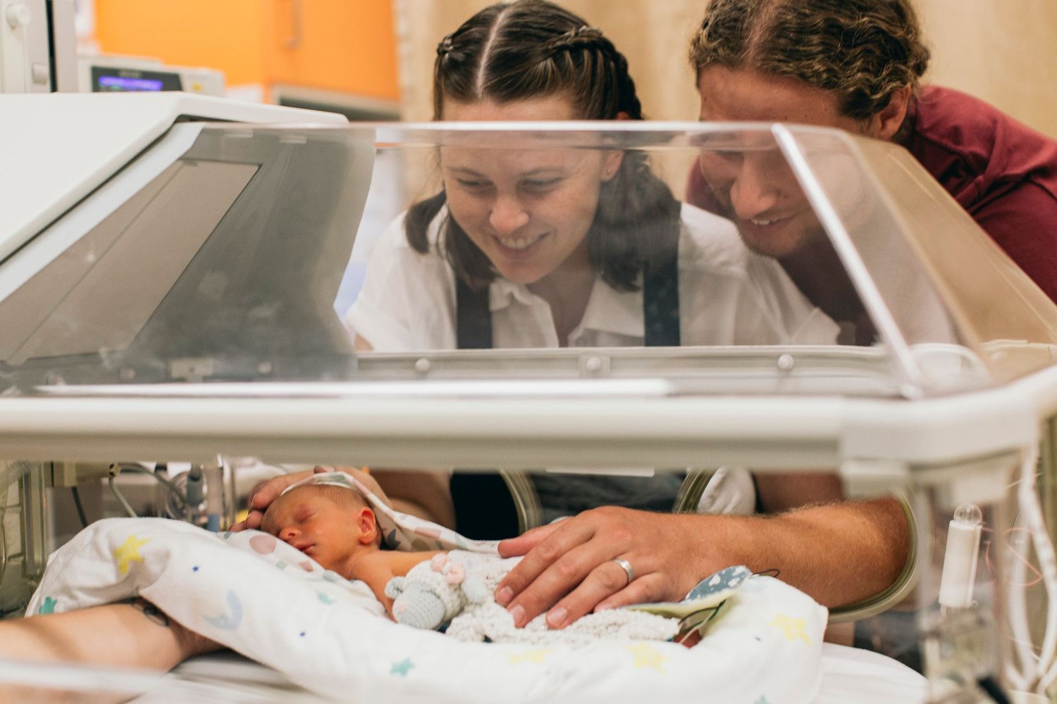 Two parents lovingly gaze at their newborn baby resting in a hospital incubator in the NICU.
