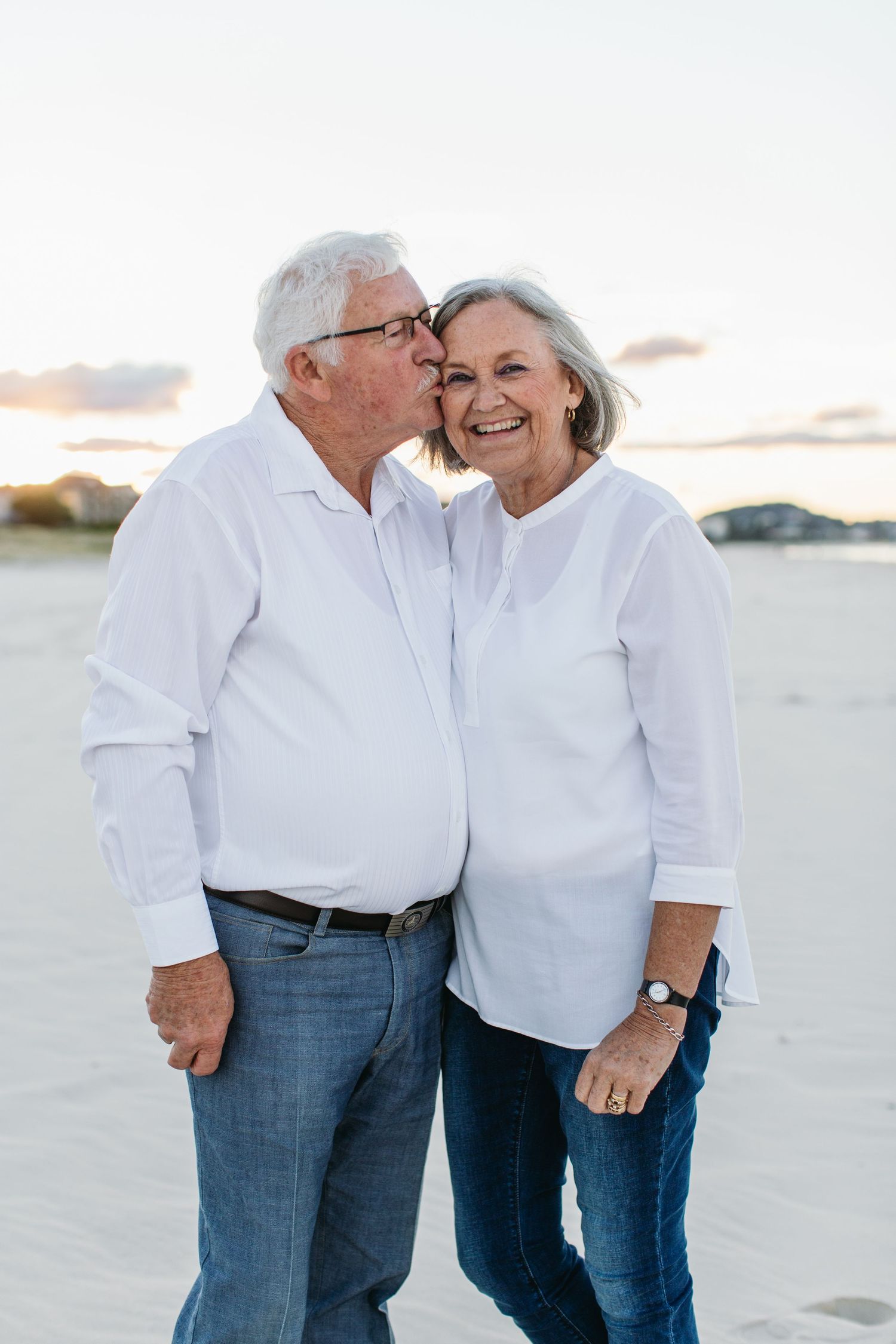 Senior couple in white shirts and jeans sharing a kiss on the beach at sunset, smiling joyfully.