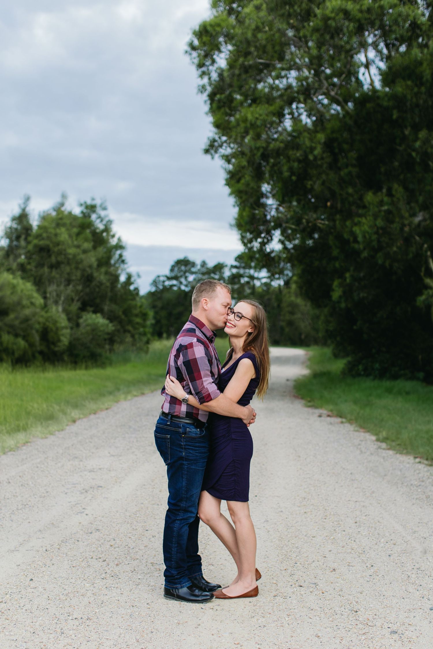 Couple embracing and smiling on a rural gravel road surrounded by lush green trees on a cloudy day.