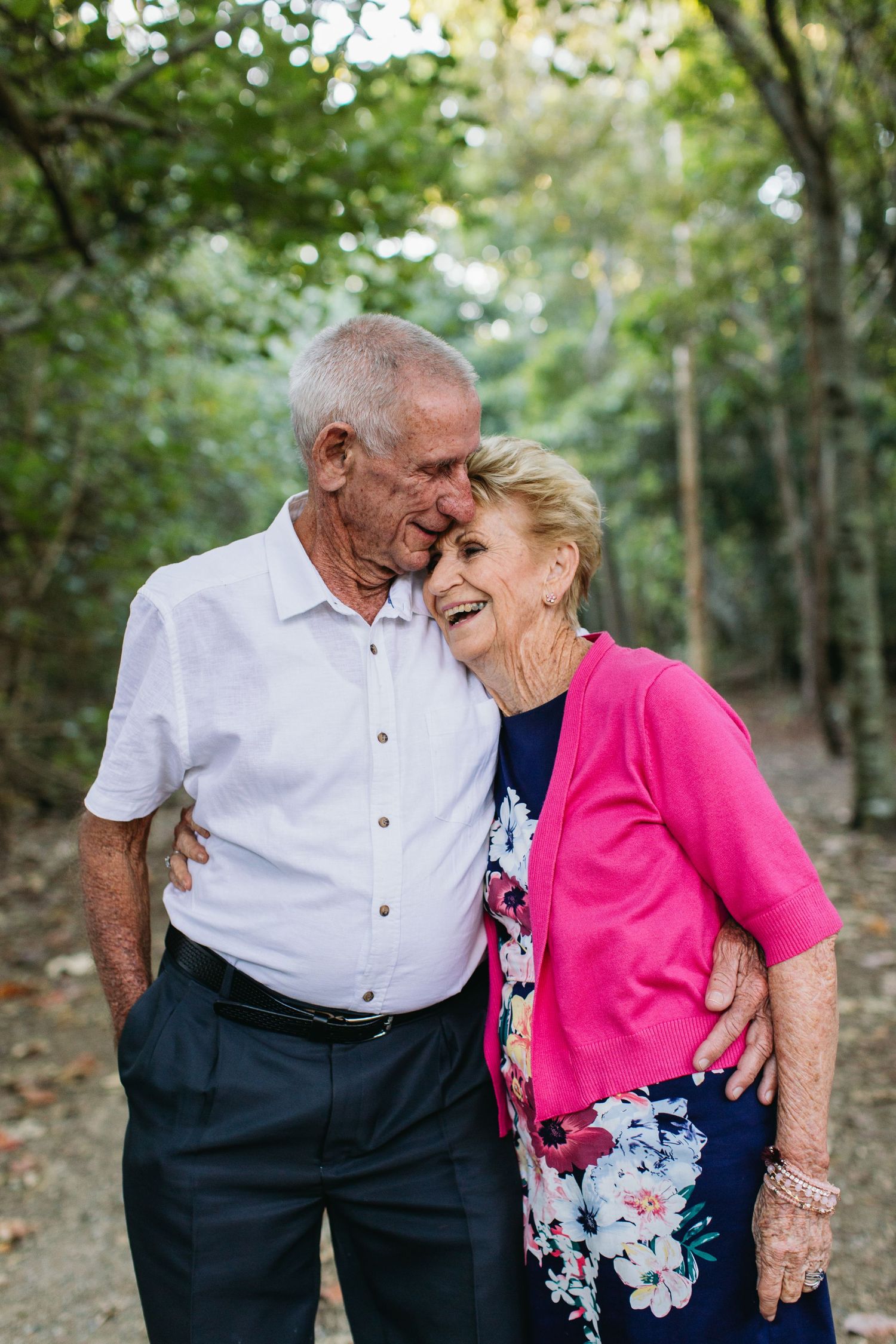 Elderly couple sharing a tender moment outdoors, woman in pink cardigan smiling as man kisses her temple.