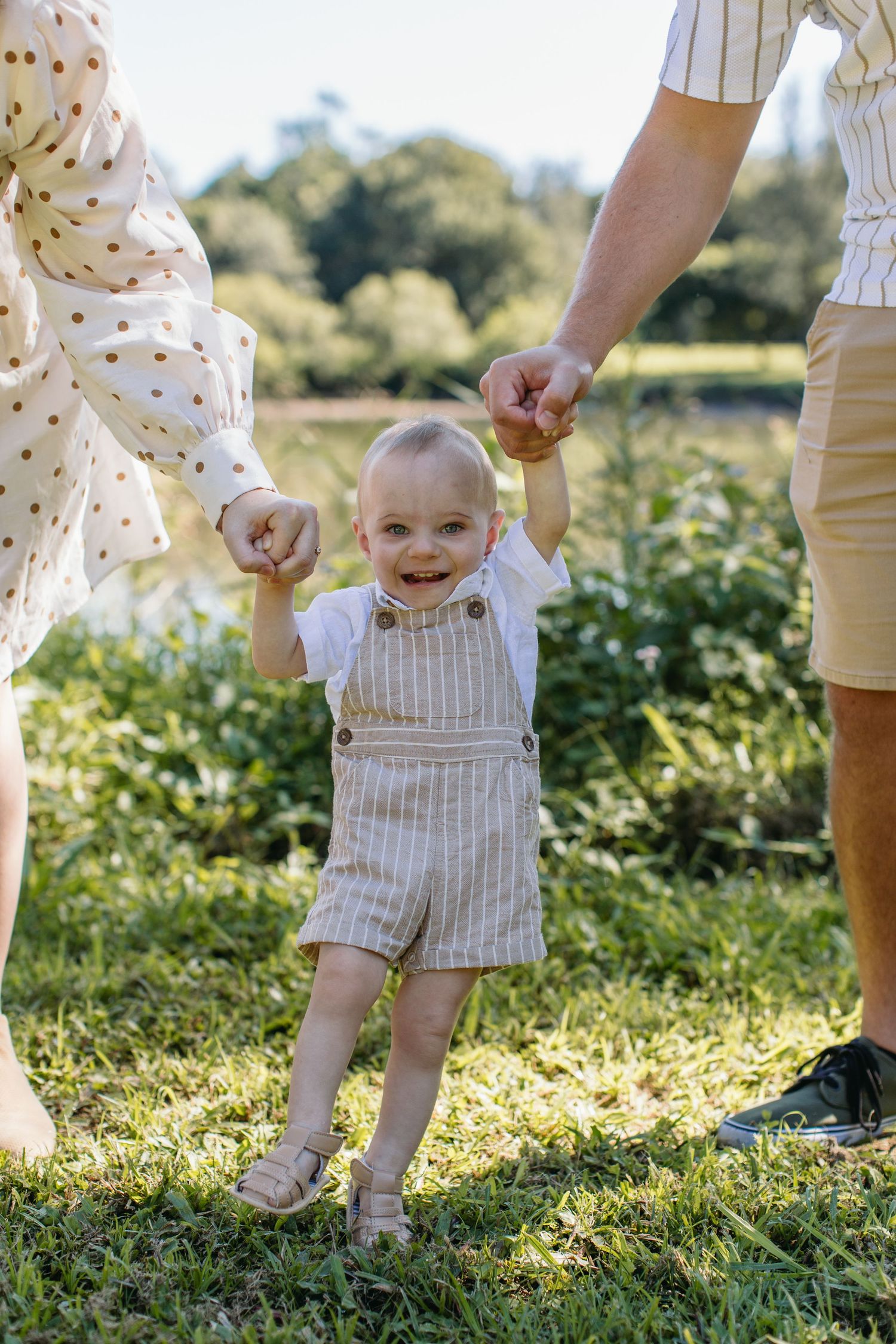 Happy baby taking first steps outdoors, held by parents hands on green grass in sunny park setting.