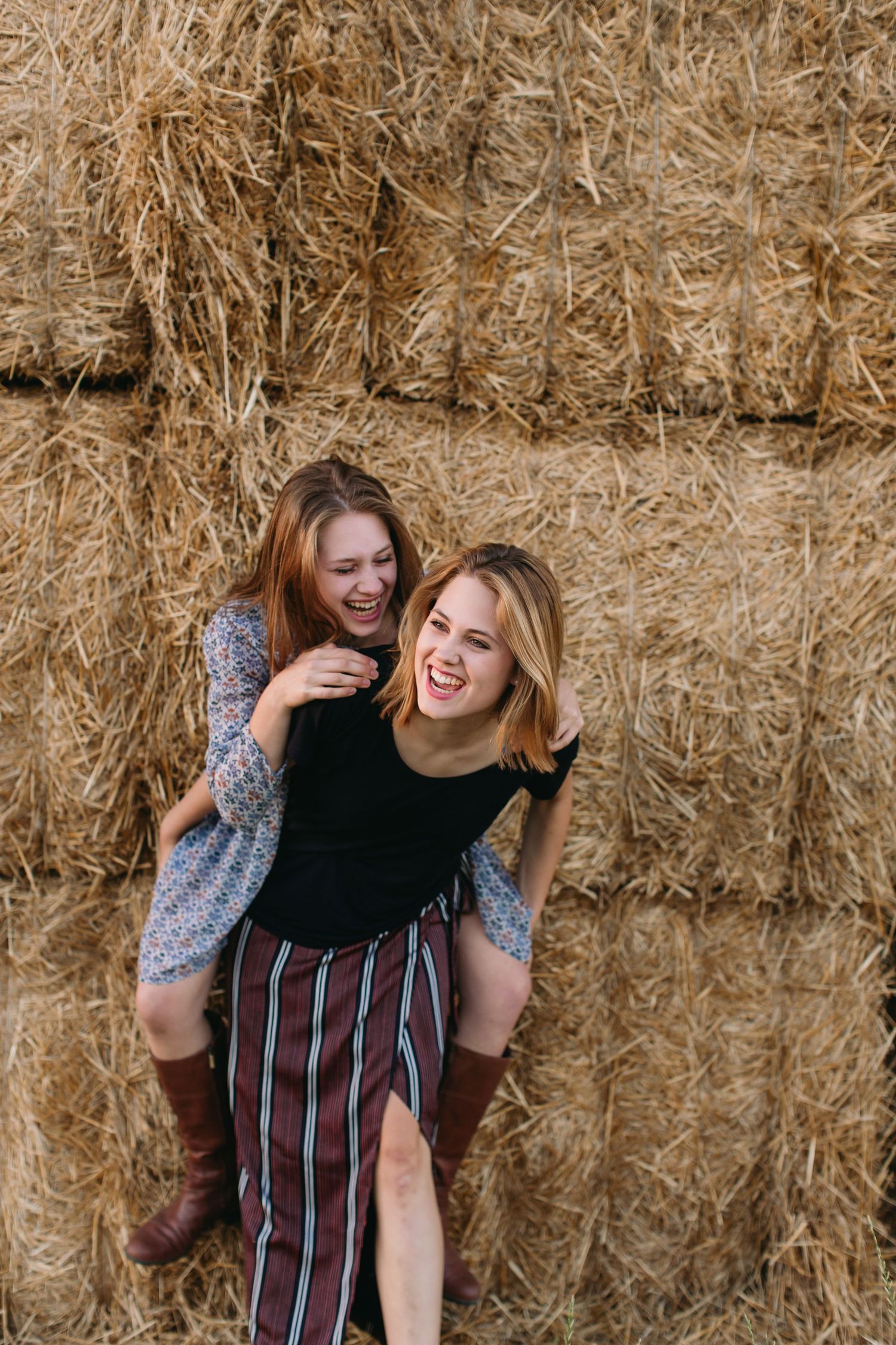 Two young women laughing and doing a piggyback ride in front of stacked hay bales on a sunny day.