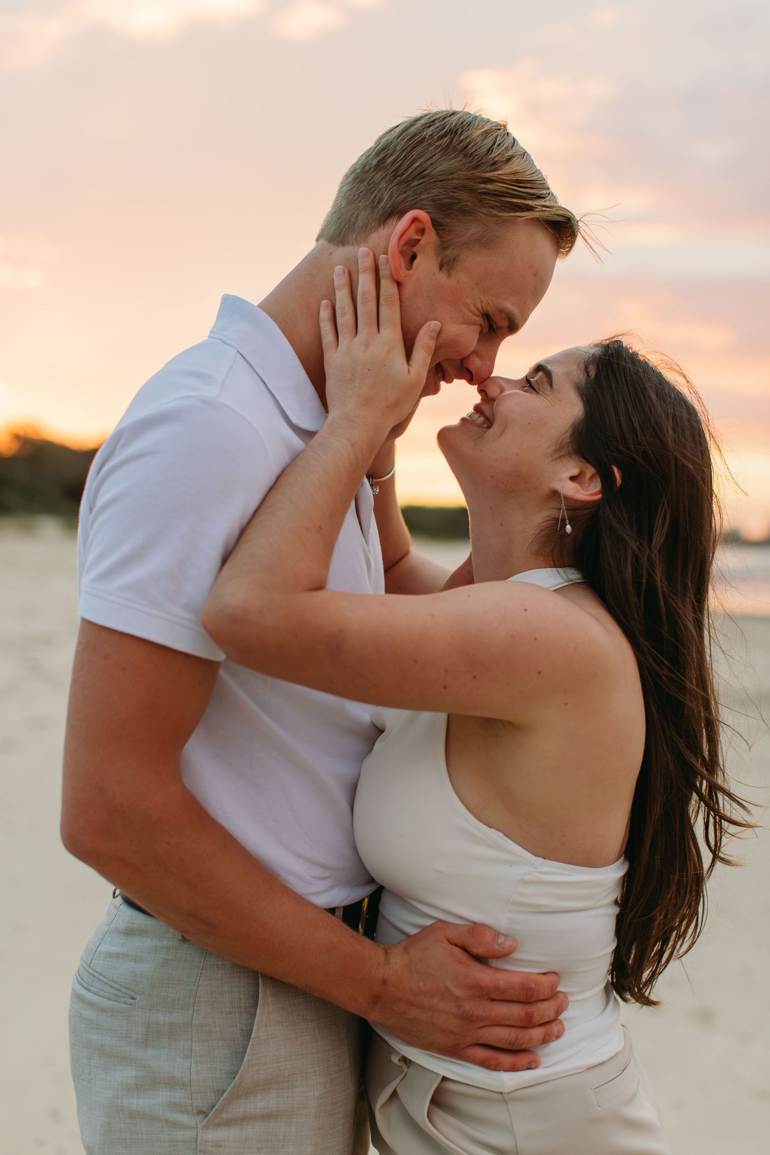 Couple sharing a romantic almost-kiss embrace on a sandy beach at sunset, woman holding man's face tenderly.