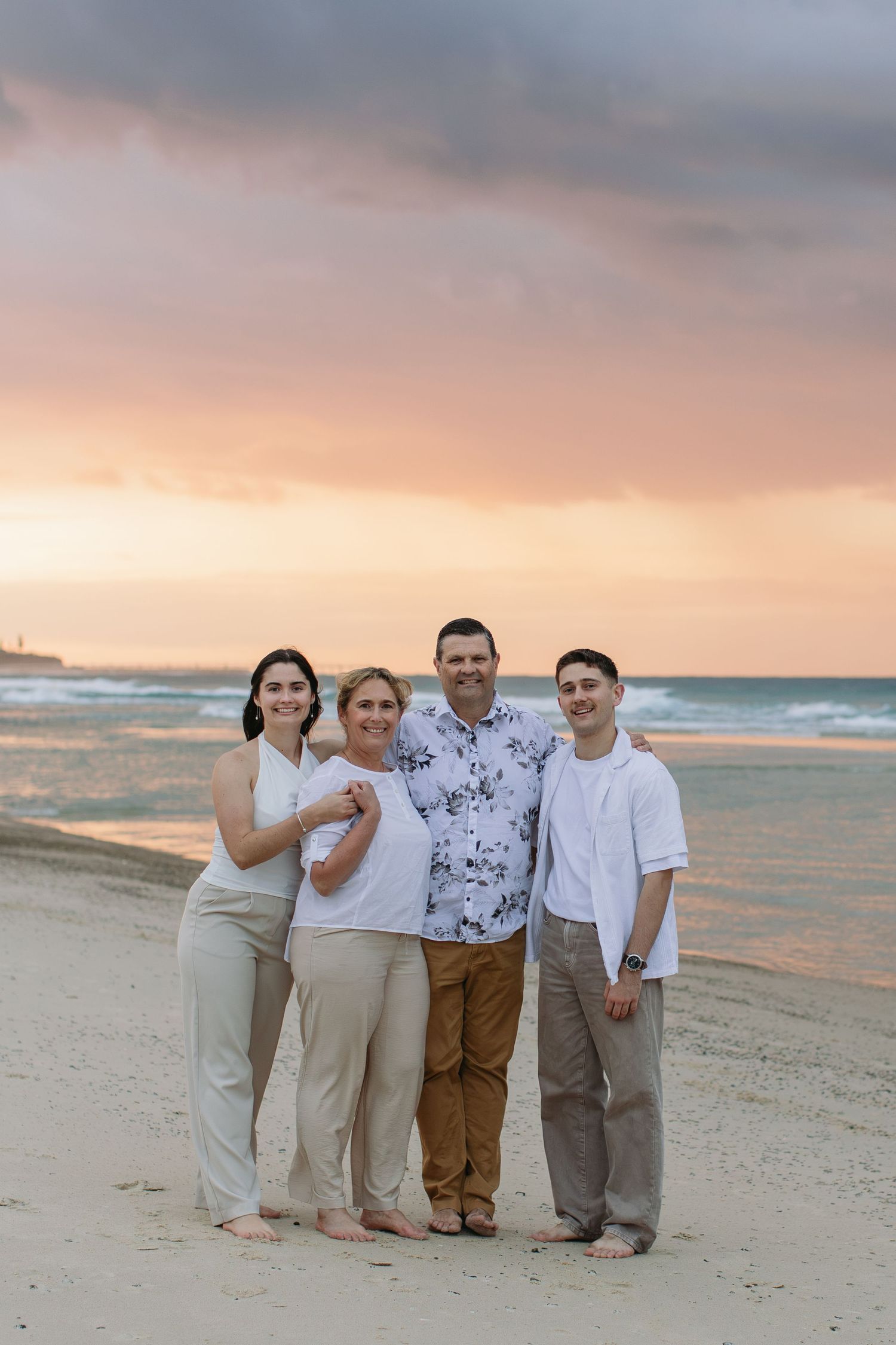 Family of four posing together on a sandy beach at sunset with colorful pink and purple sky over the ocean.