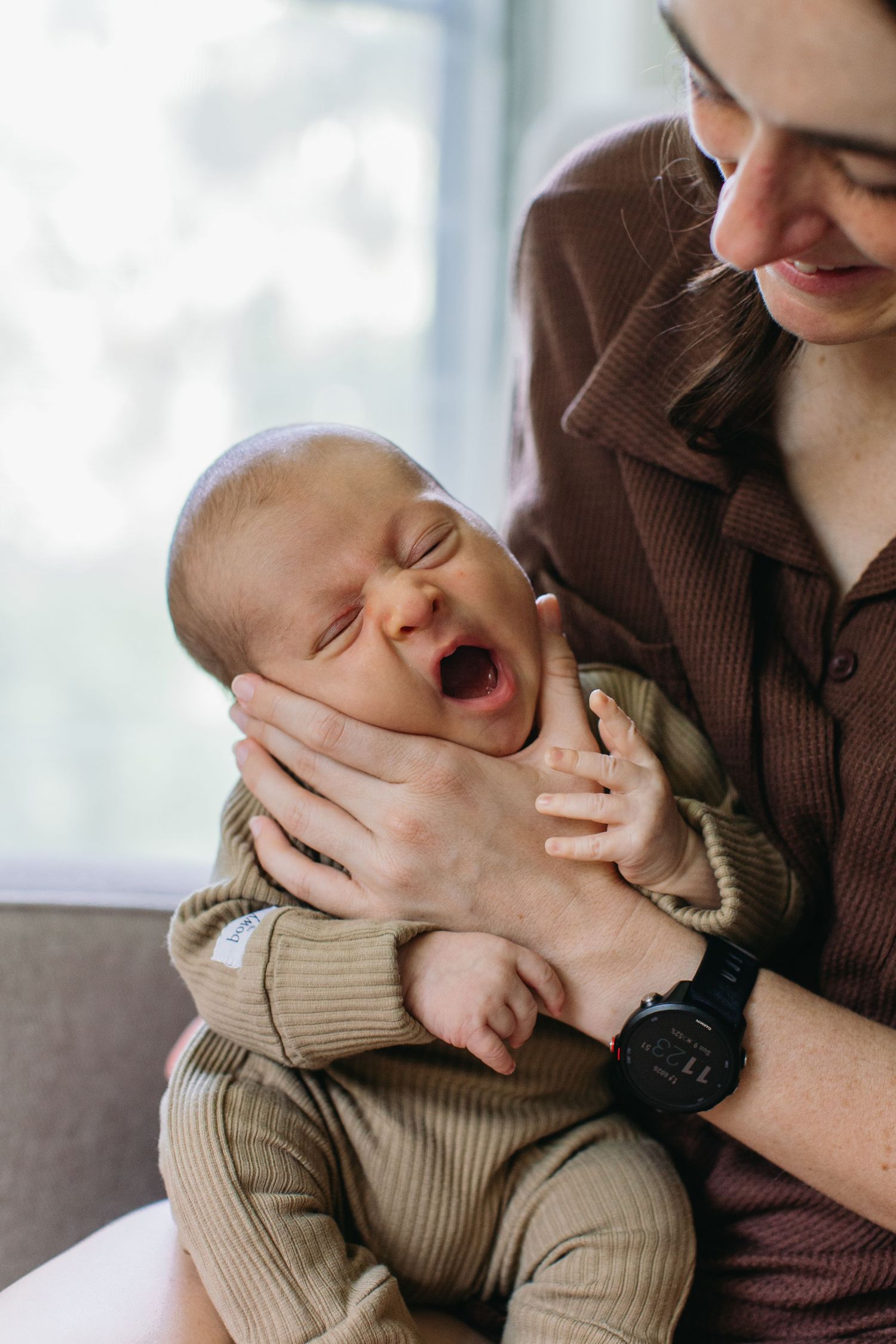 Mother holding a yawning newborn baby dressed in olive green outfit indoors.