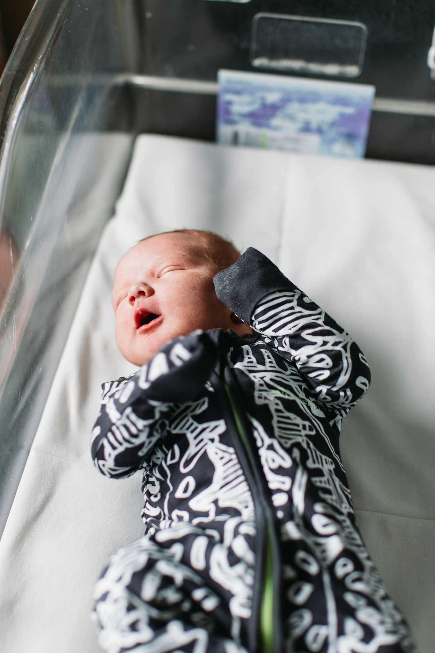 Crying newborn baby in black and white patterned onesie lying in hospital bassinet.