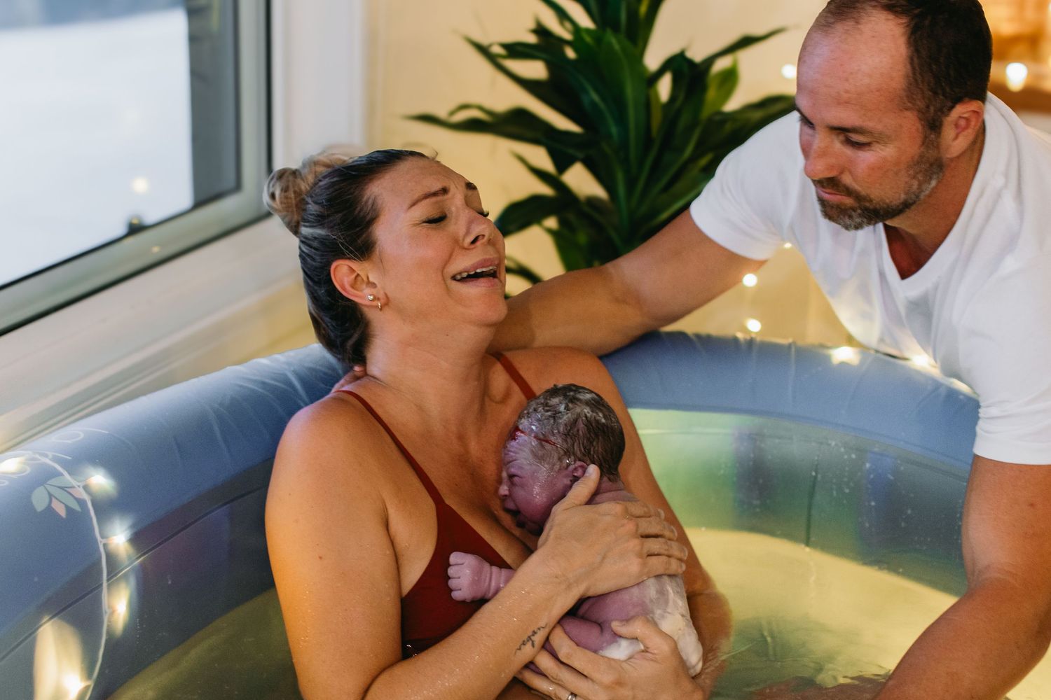 Joyful mother holding newborn baby in water birth pool while partner supports her during delivery.