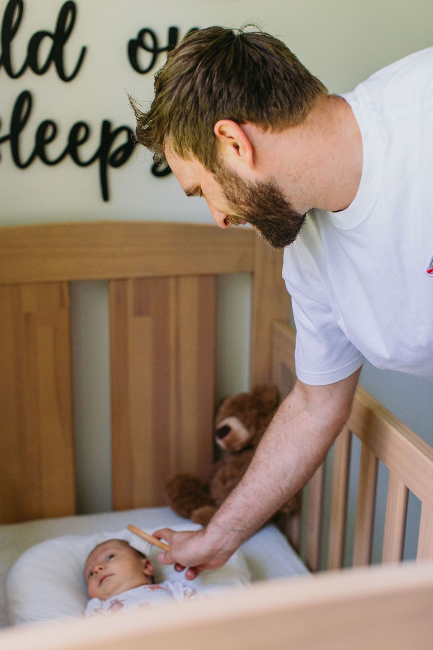 Bearded father in white t-shirt gently placing newborn baby into wooden crib with teddy bear during in-home newborn session.