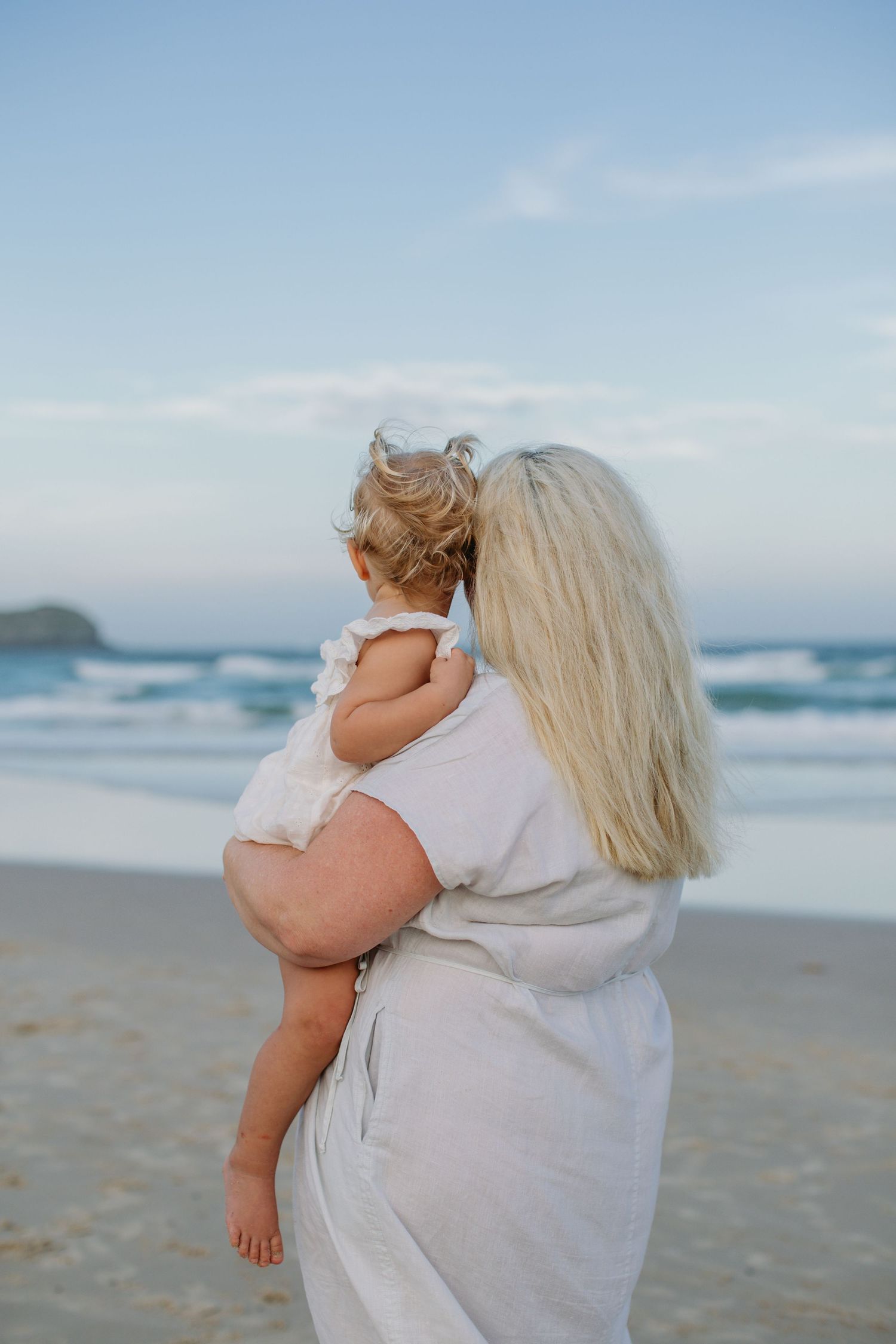 A blonde woman holding a toddler girl on a sandy beach, both facing the ocean waves under a soft blue sky.