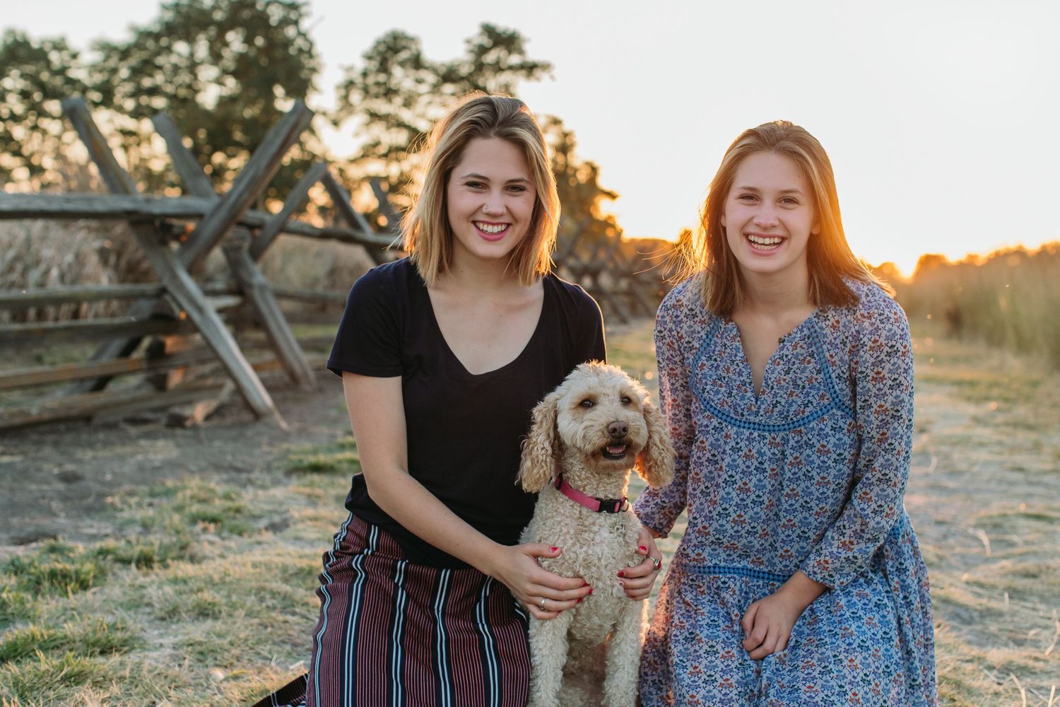Two smiling women pose outdoors at sunset with a fluffy golden doodle dog on a farm.