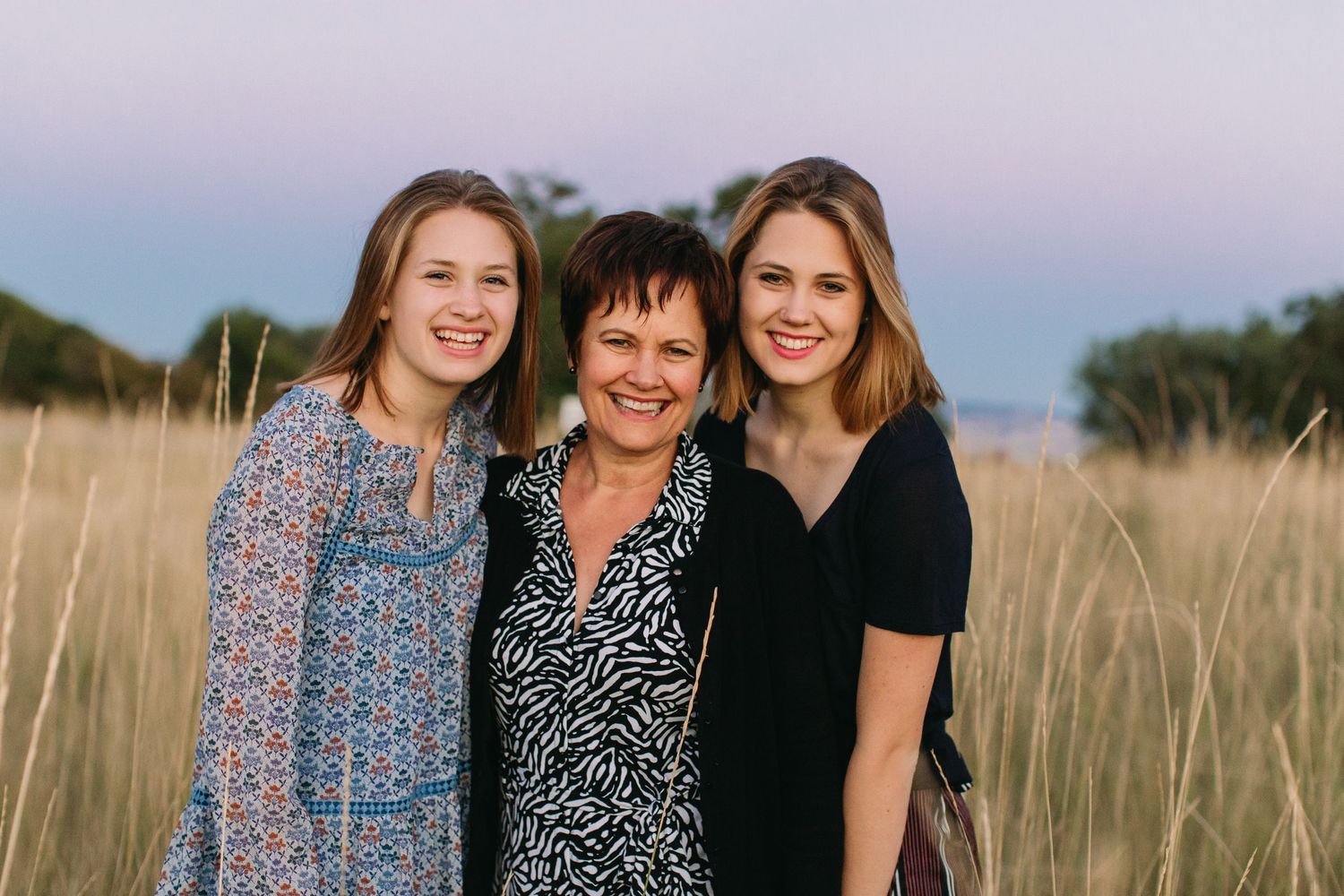 Three smiling women pose together in a golden grass field at sunset, wearing casual dresses in a warm outdoor setting.