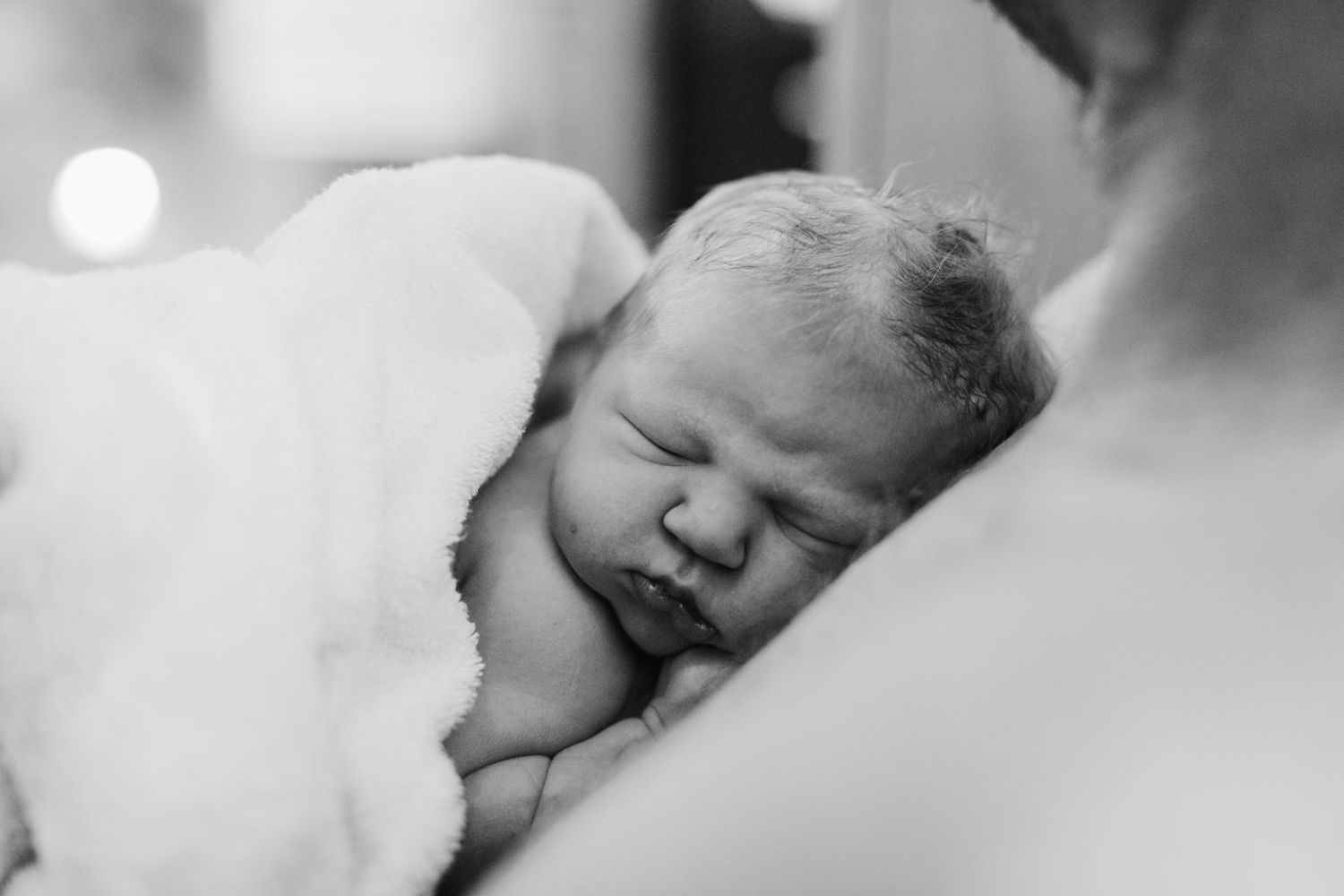 Black and white photo of a sleeping newborn baby wrapped in a soft white blanket cradled in a parents arms.