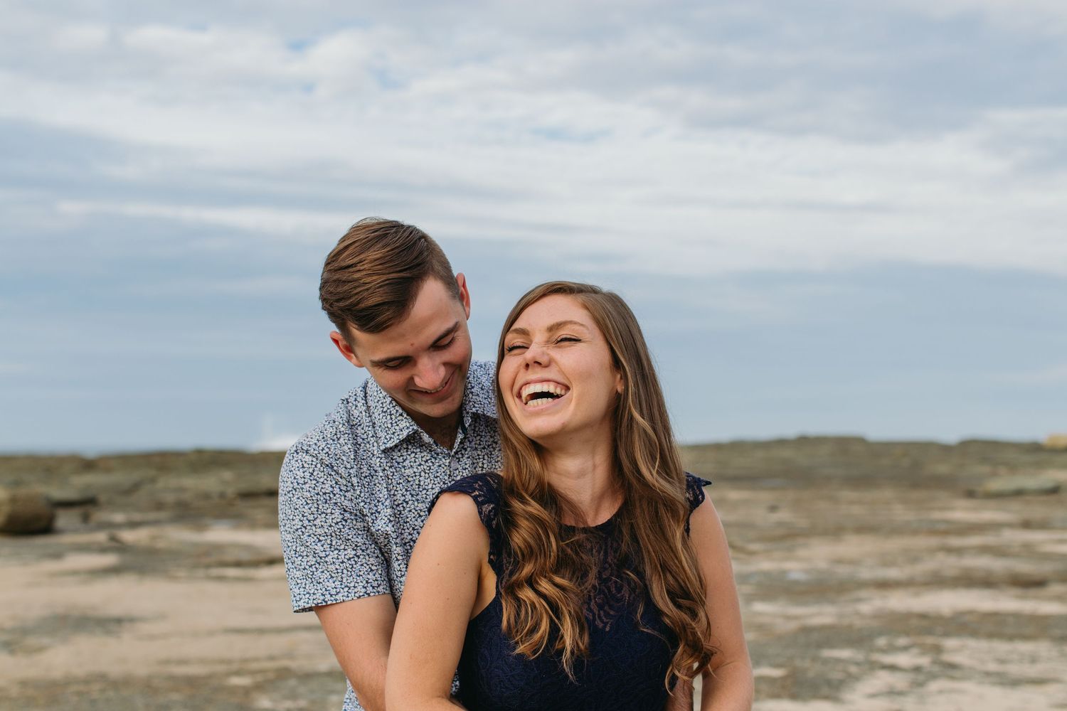 Happy couple laughing together on a rocky beach with cloudy sky backdrop during an outdoor engagement photo session.