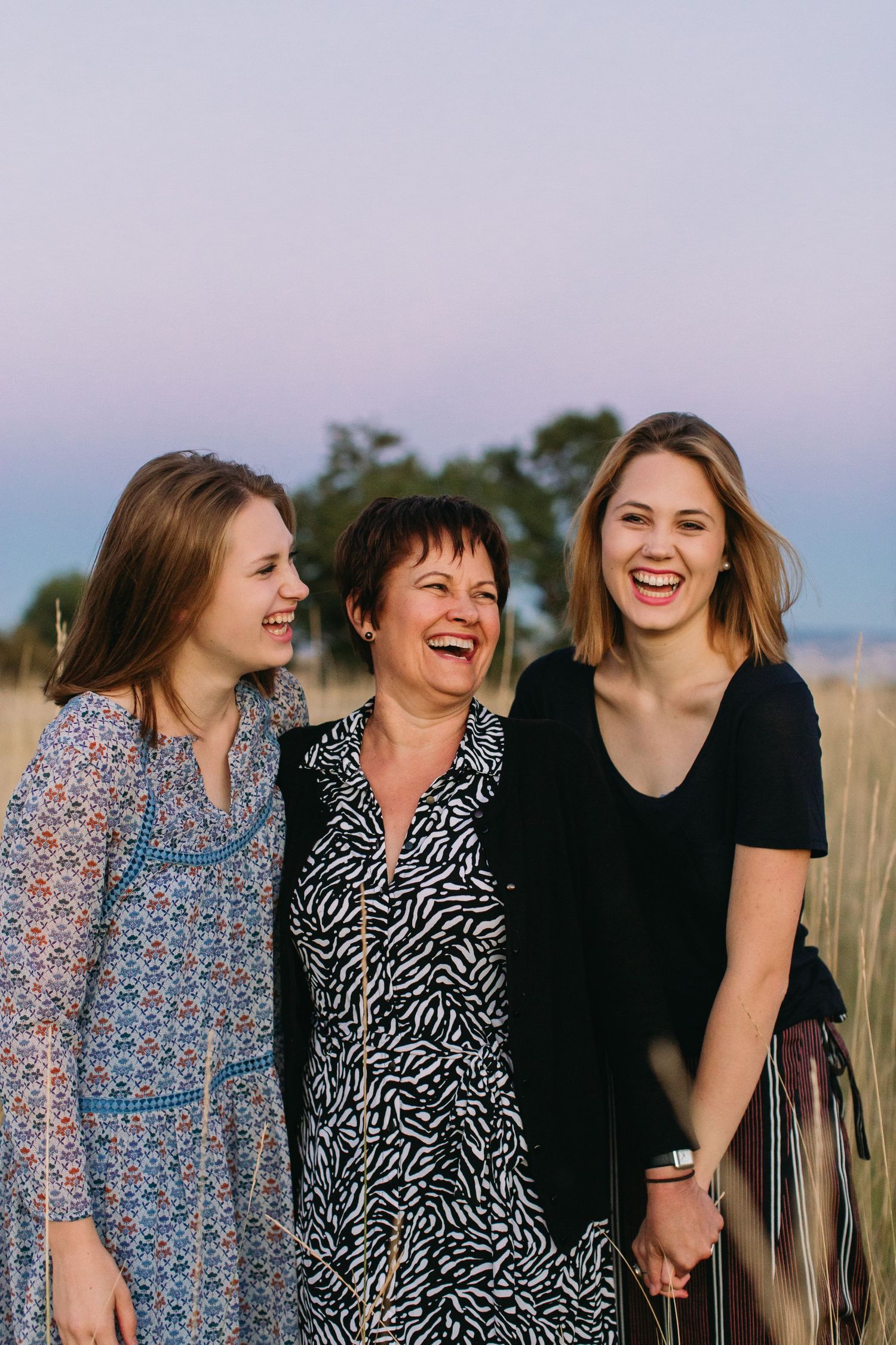Three women laughing together outdoors in a golden field at dusk, wearing casual dresses in a warm family portrait.