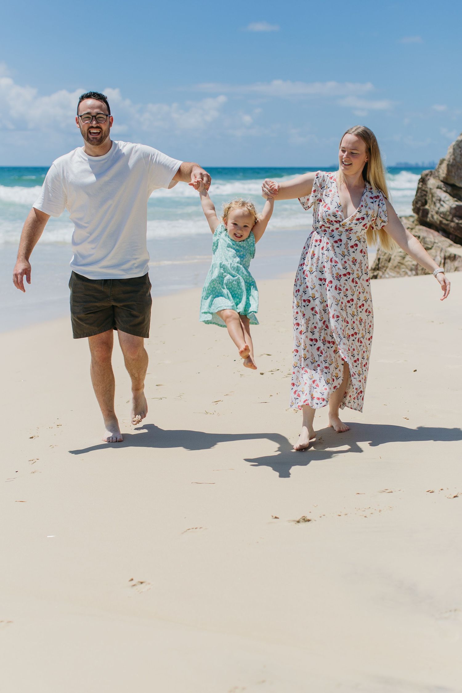 A happy family swings their young daughter on a sunny beach with turquoise waves and rocky shores in the background.