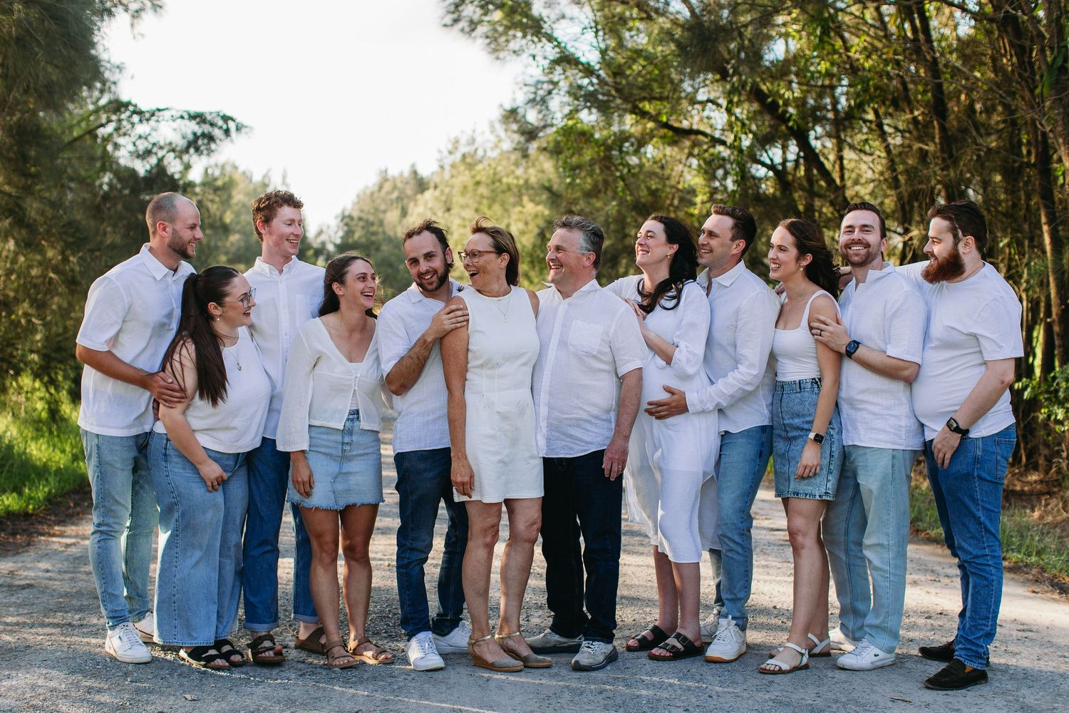 Large family group posing outdoors in matching white tops and jeans, smiling and laughing together on a tree-lined path.