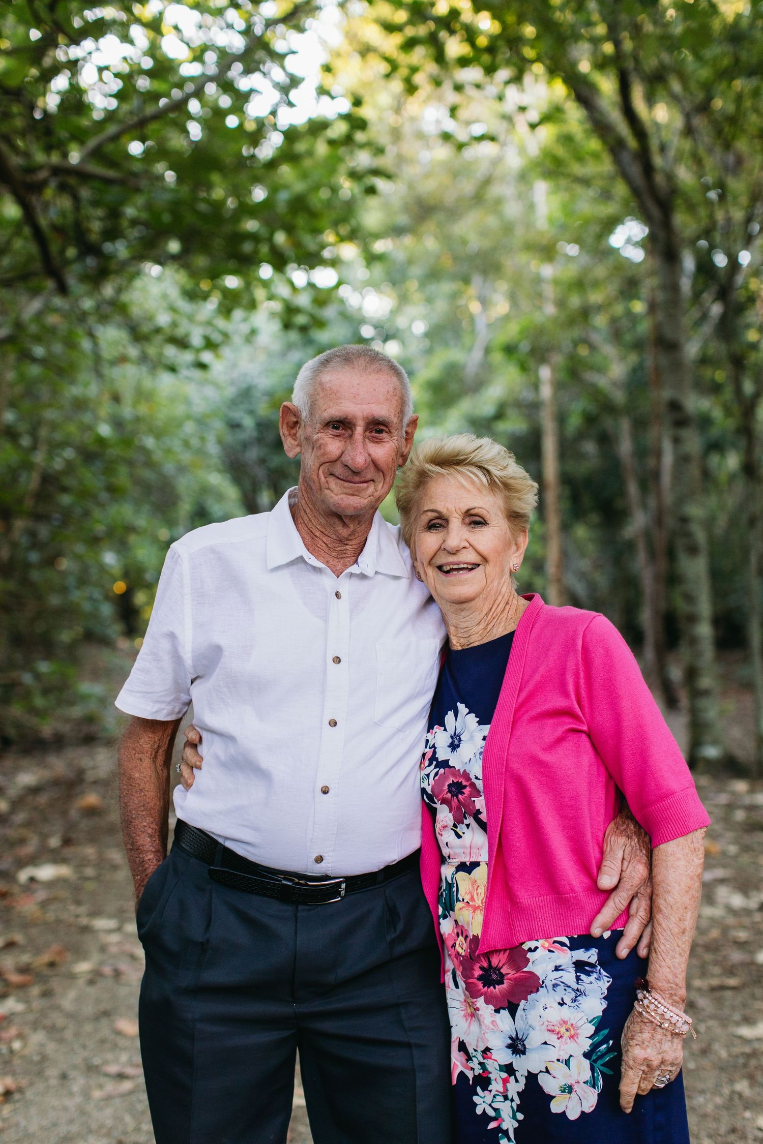 Smiling elderly couple posing together outdoors in a lush green tree-lined path, woman in pink cardigan and floral dress.