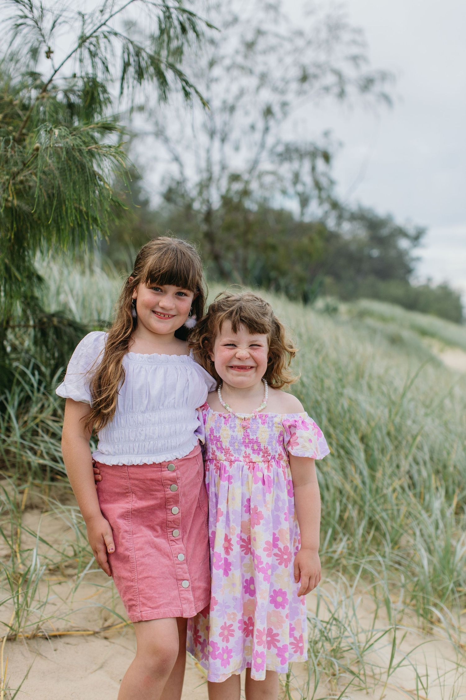Two smiling young sisters posing together on a sandy beach path surrounded by coastal greenery.