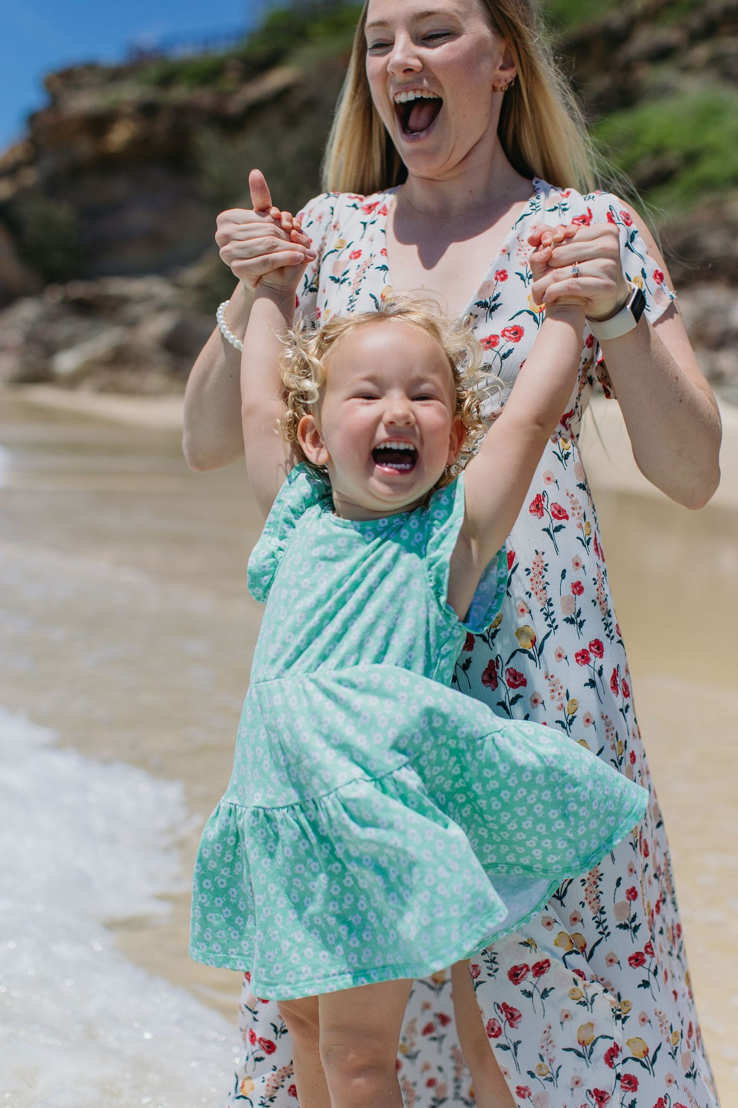 Excited mother spinning her laughing toddler daughter in a teal dress on a sunny beach.