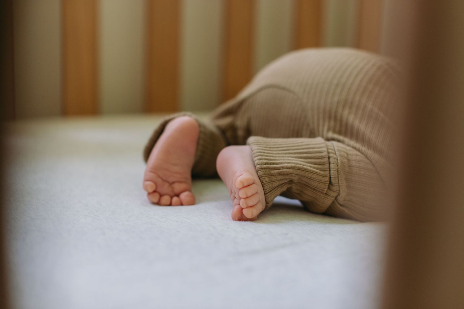 Close-up of baby feet in a brown ribbed onesie lying in a white crib.