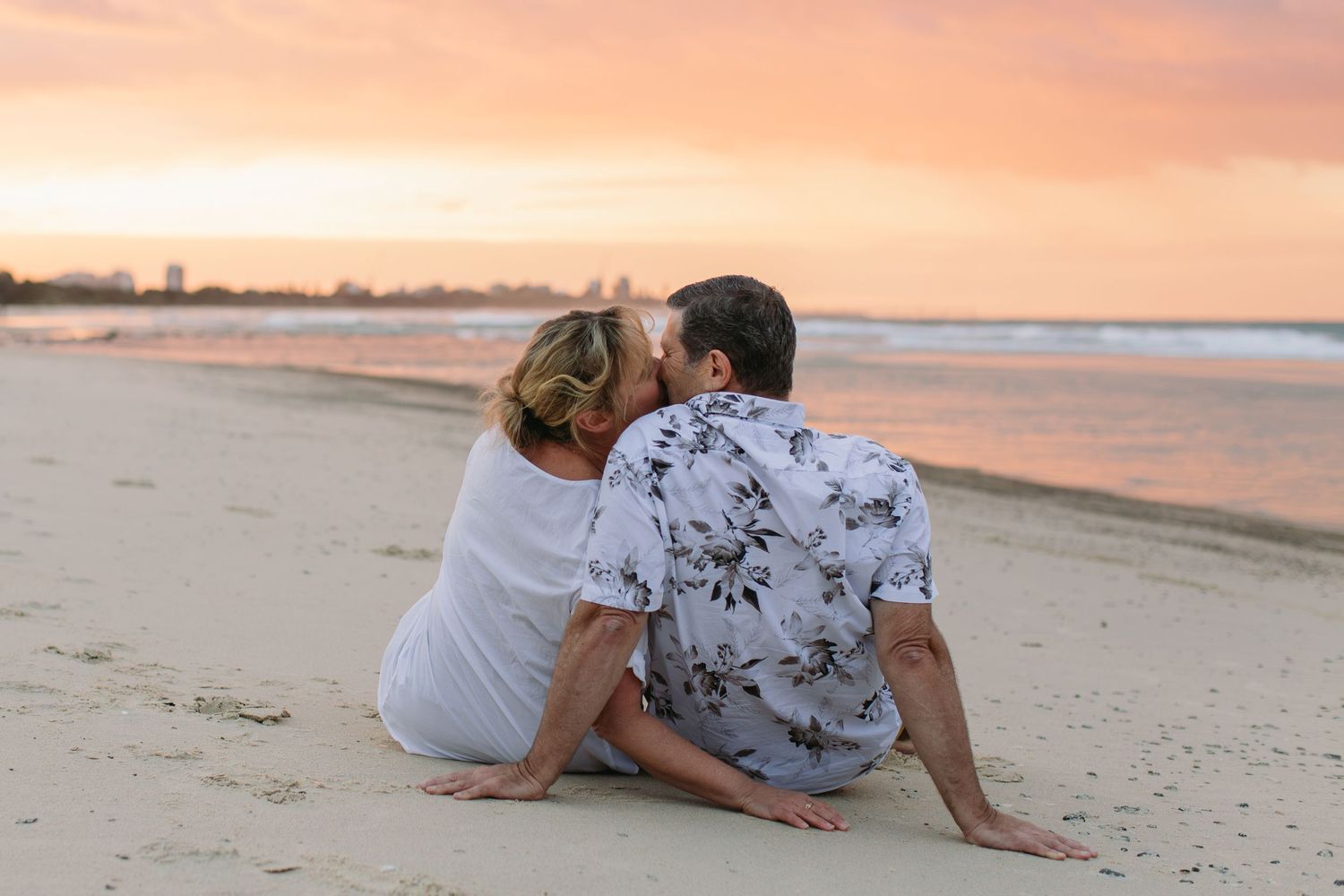A couple sits on a sandy beach sharing a kiss while watching a stunning pink and orange sunset over the ocean.