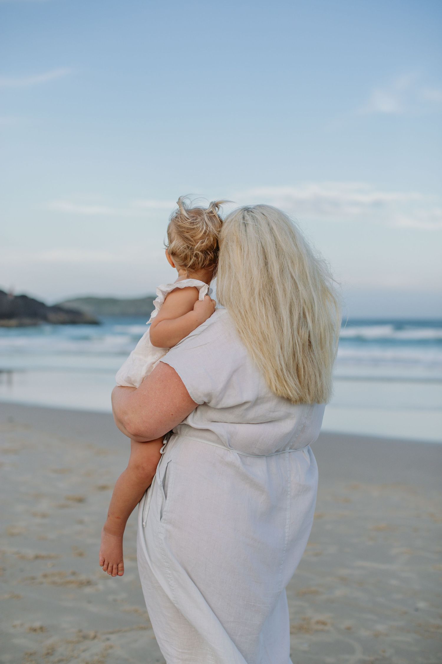 Blonde mother holding toddler on sandy beach with ocean waves in background.