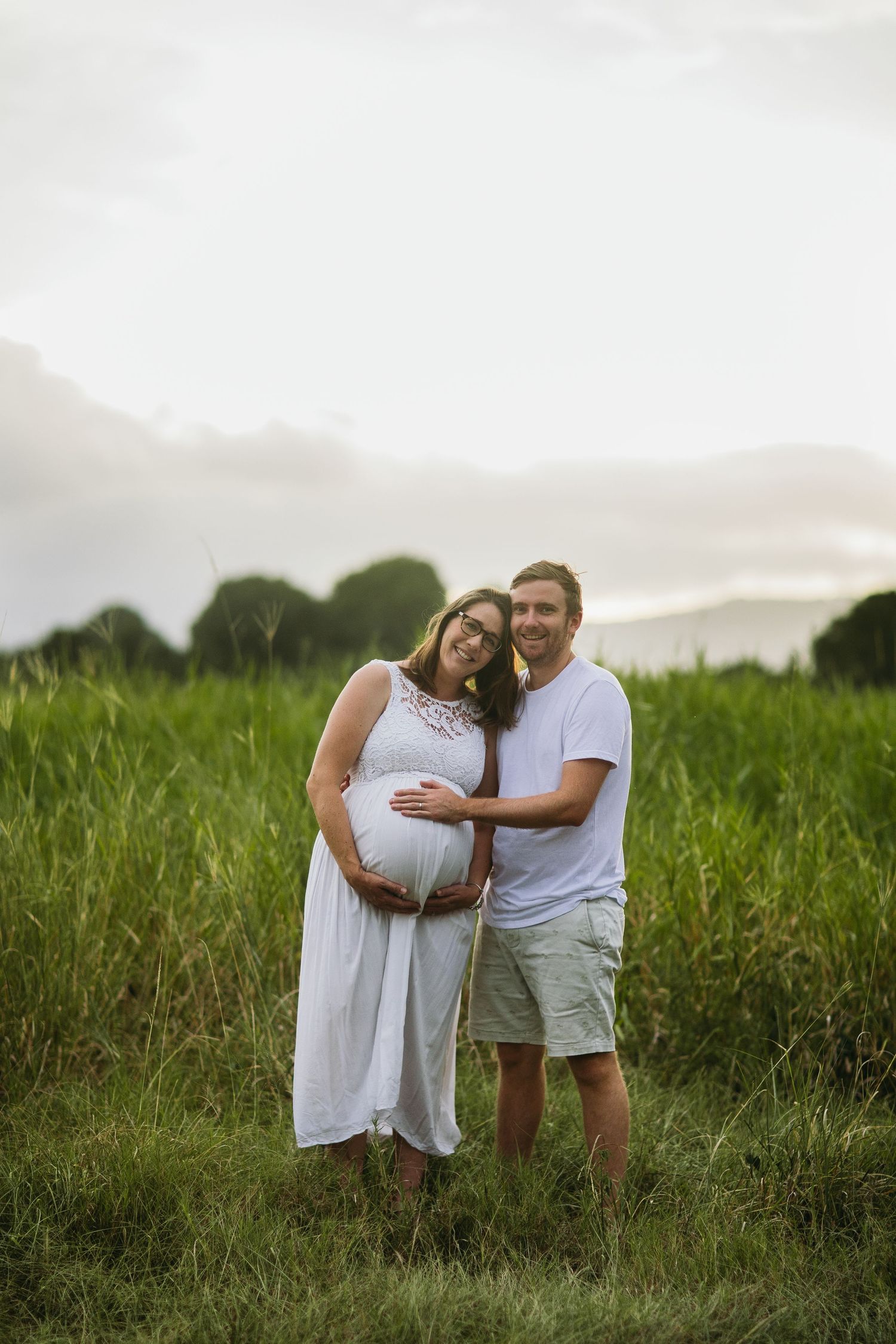 Pregnant couple posing in a green field, woman in white dress cradling baby bump, man in white shirt embracing her from behind.