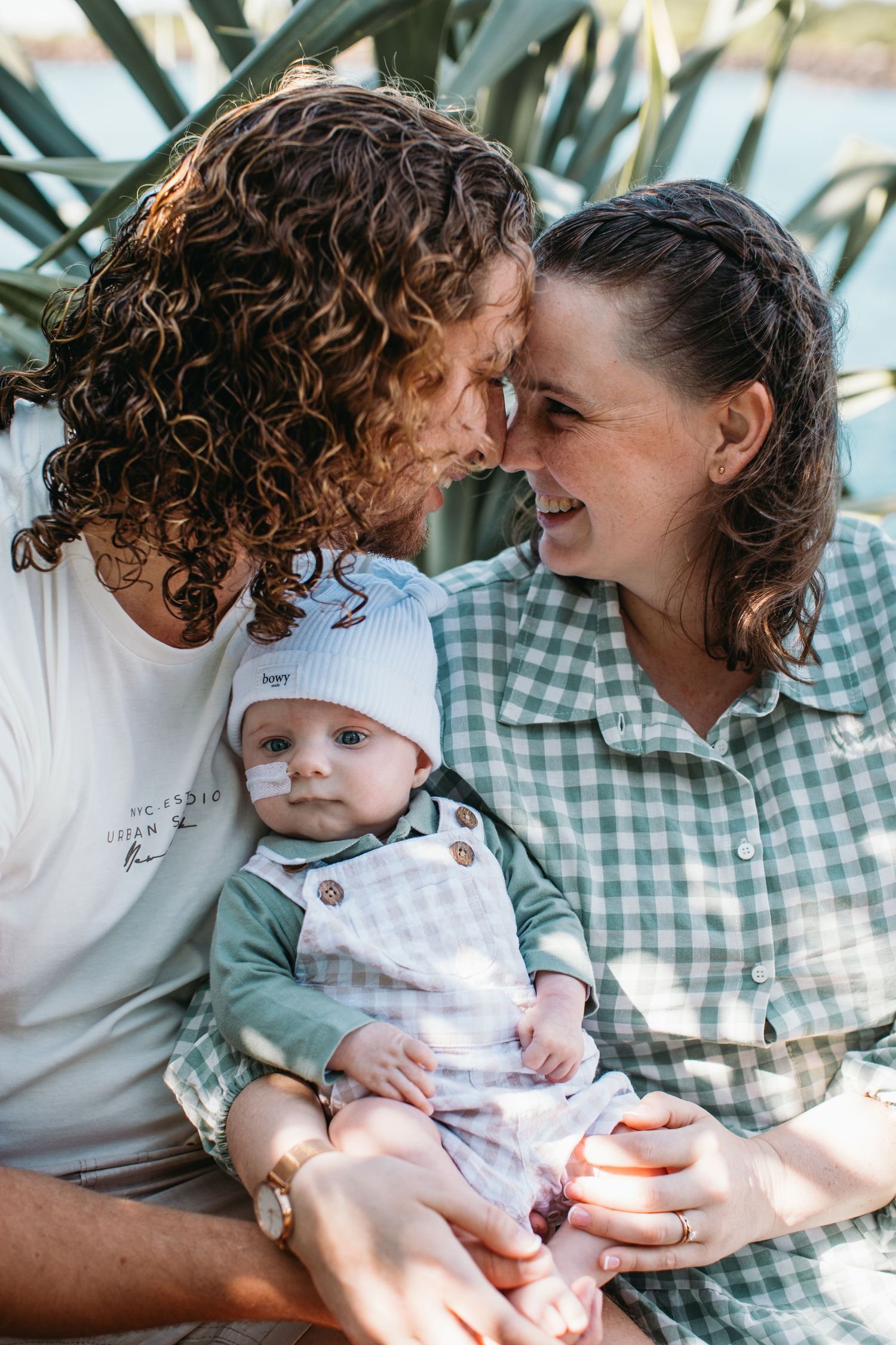A couple with curly hair touch foreheads while holding their baby in a hat outdoors near tropical plants.