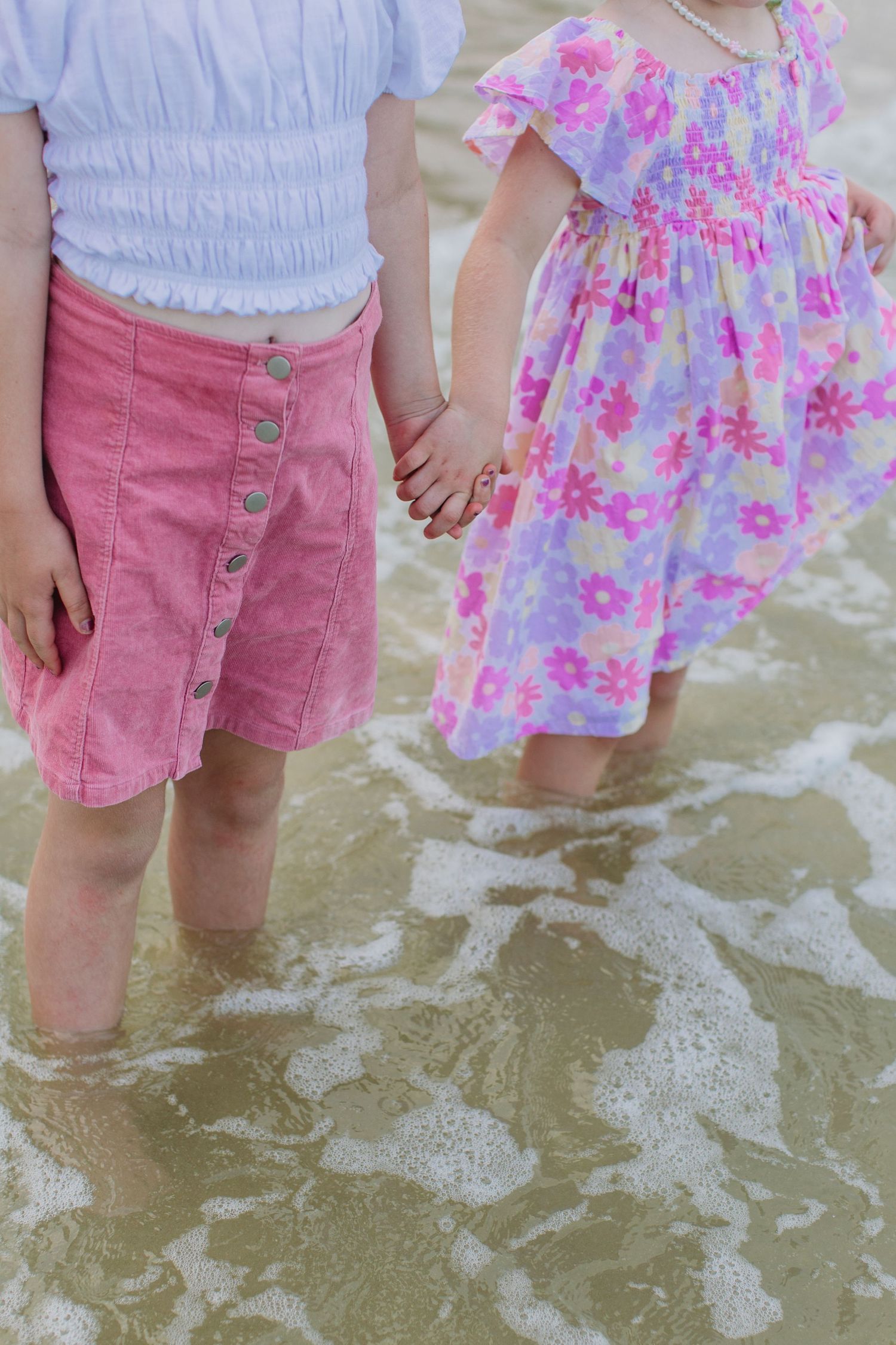 Two young girls in pink outfits hold hands while standing in shallow ocean waves at the beach.