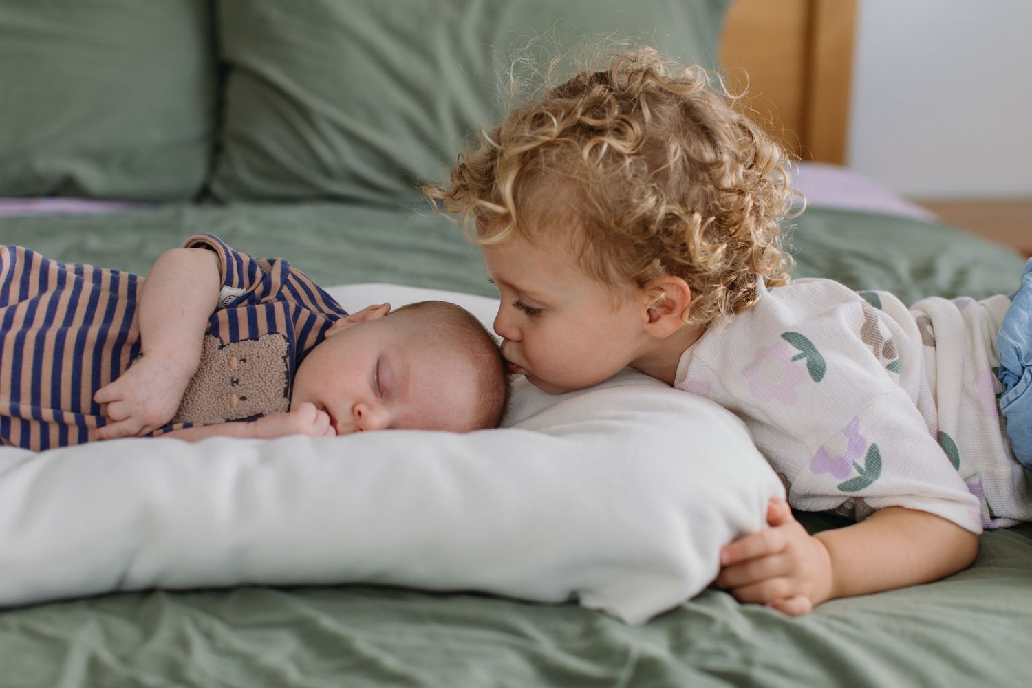 Curly-haired toddler gently kissing sleeping newborn baby sibling on a green bed with white pillow.