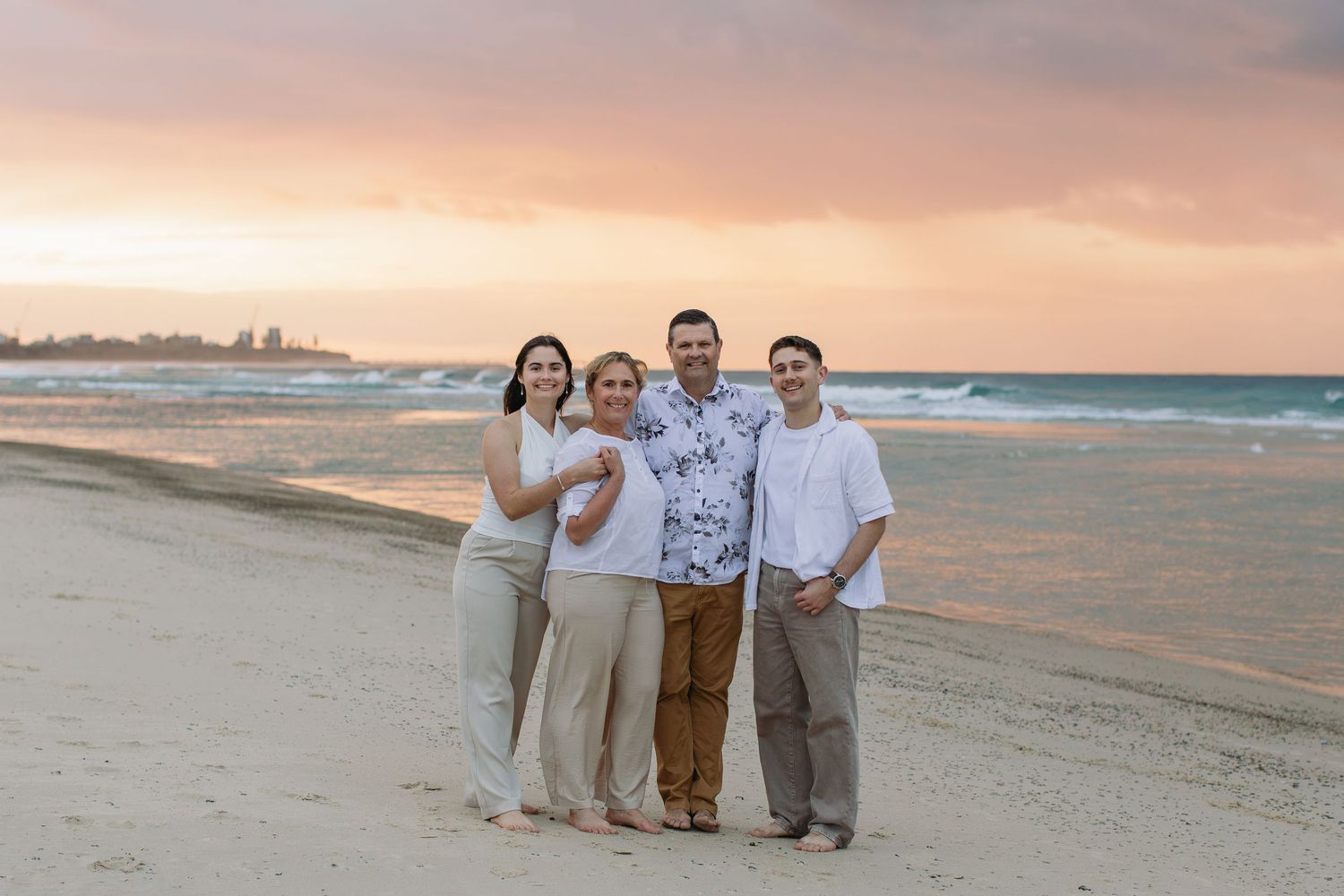 Family of four poses together on a sandy beach at sunset with colorful pink and orange skies over the ocean.