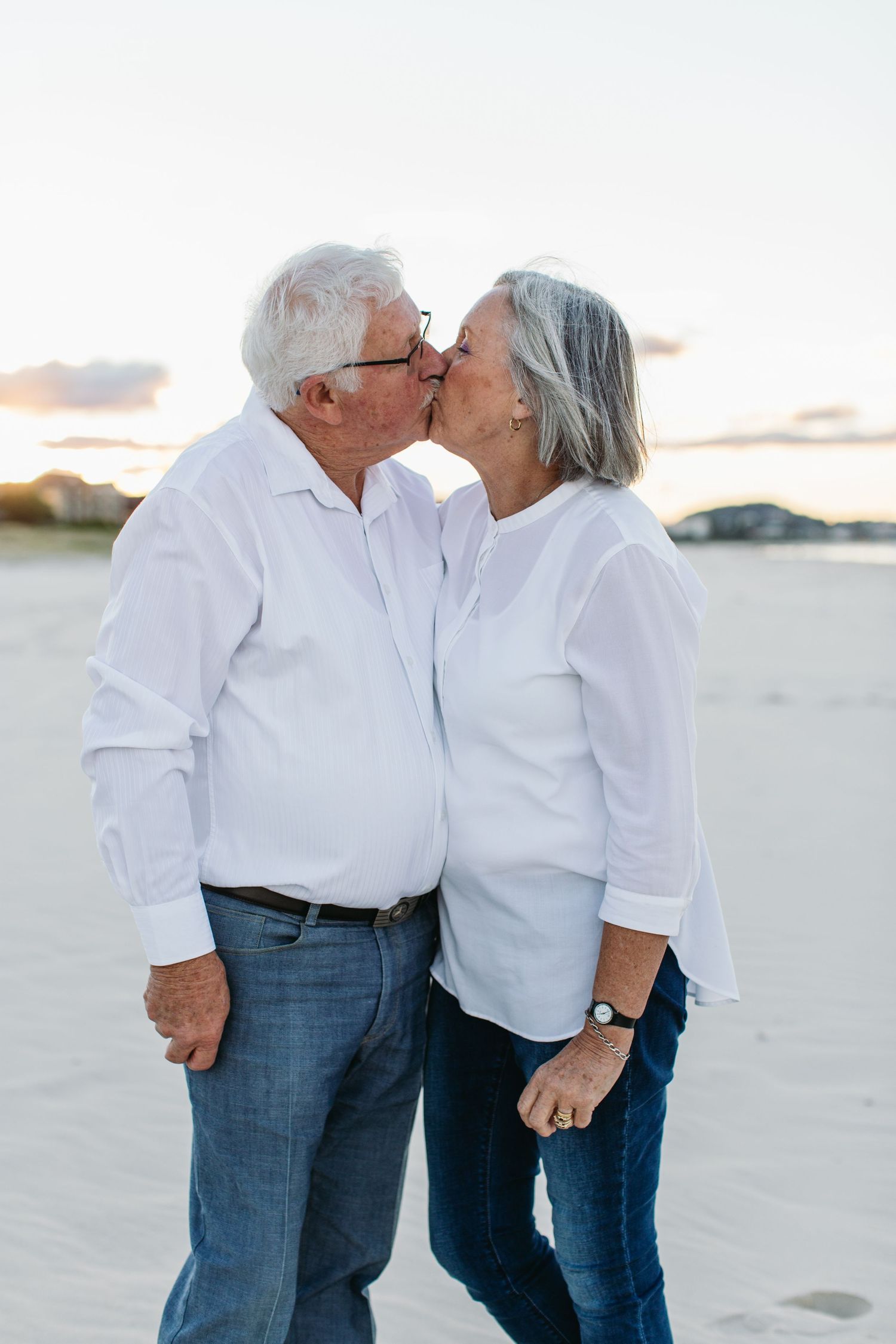 Elderly couple sharing a romantic kiss on a sandy beach at sunset, both wearing matching white shirts and blue jeans.
