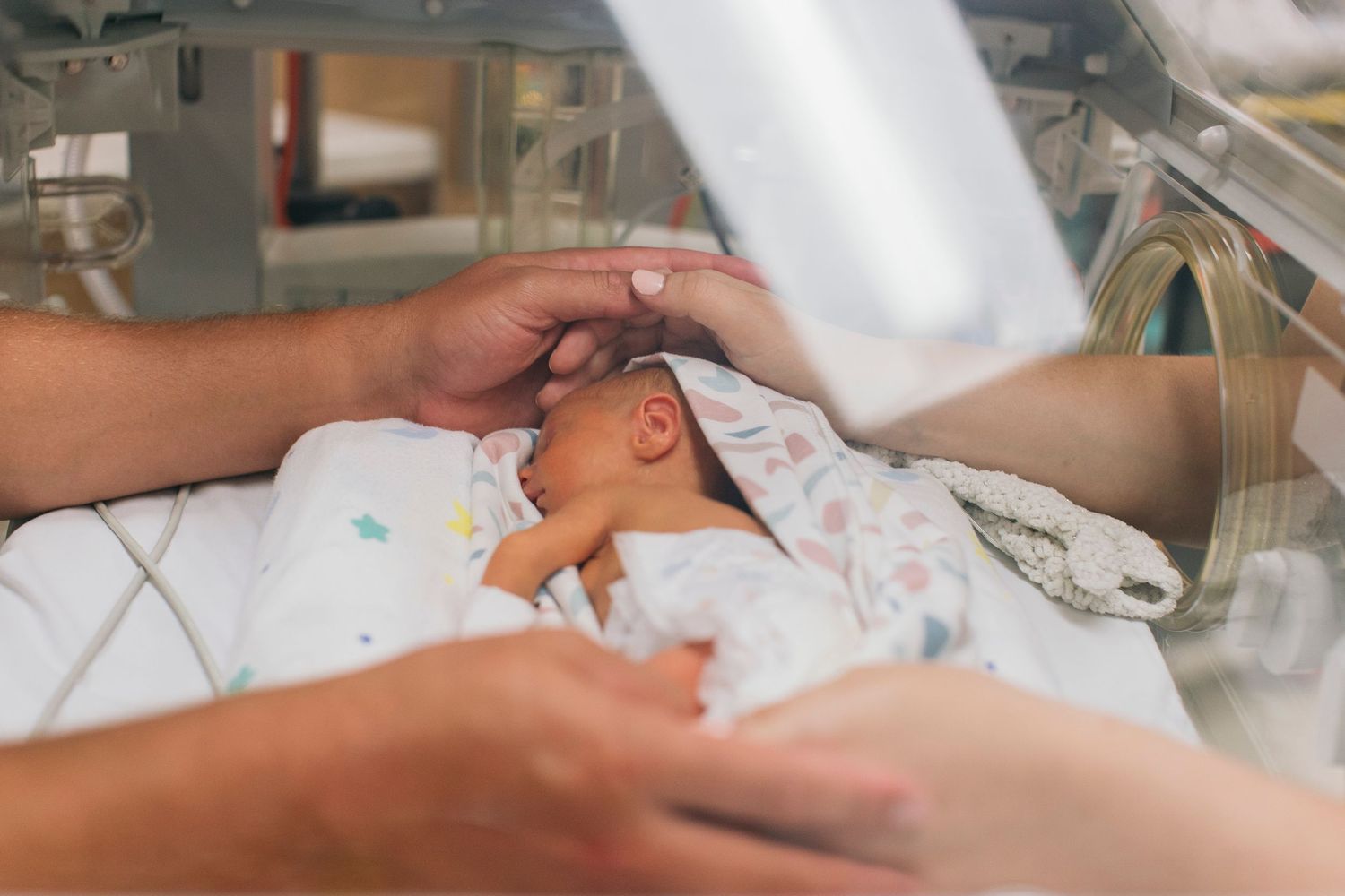 Premature newborn baby resting in a hospital incubator while parents gently touch and comfort their infant.