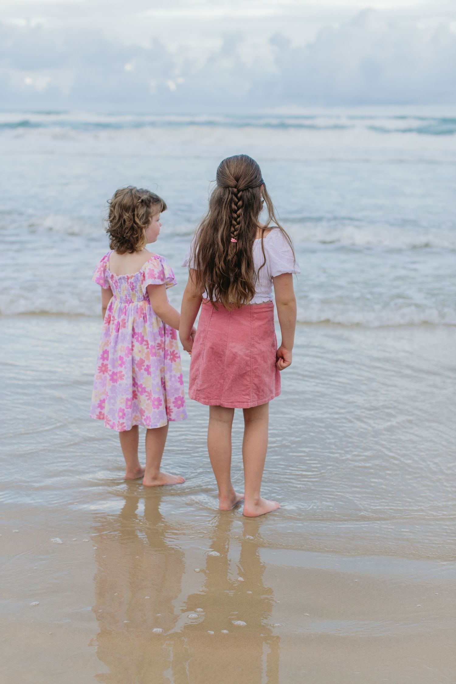 Two young girls in pink dresses hold hands while looking out at ocean waves from the shoreline.