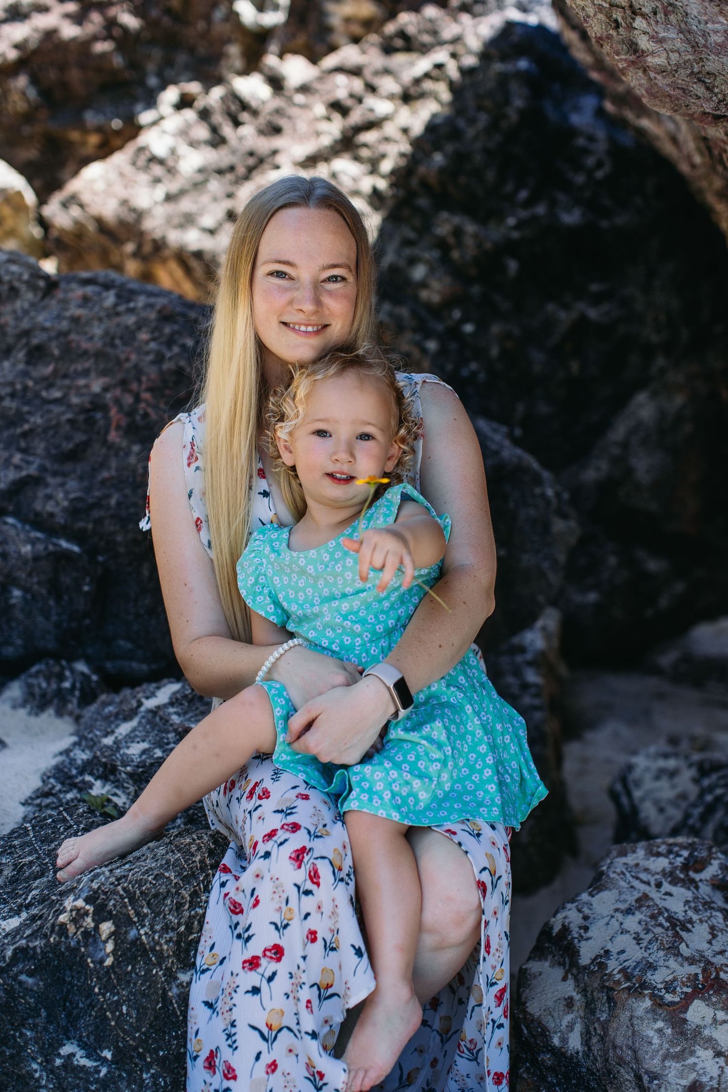 Smiling blonde woman sitting on rocks hugging a curly-haired toddler in a turquoise dress.