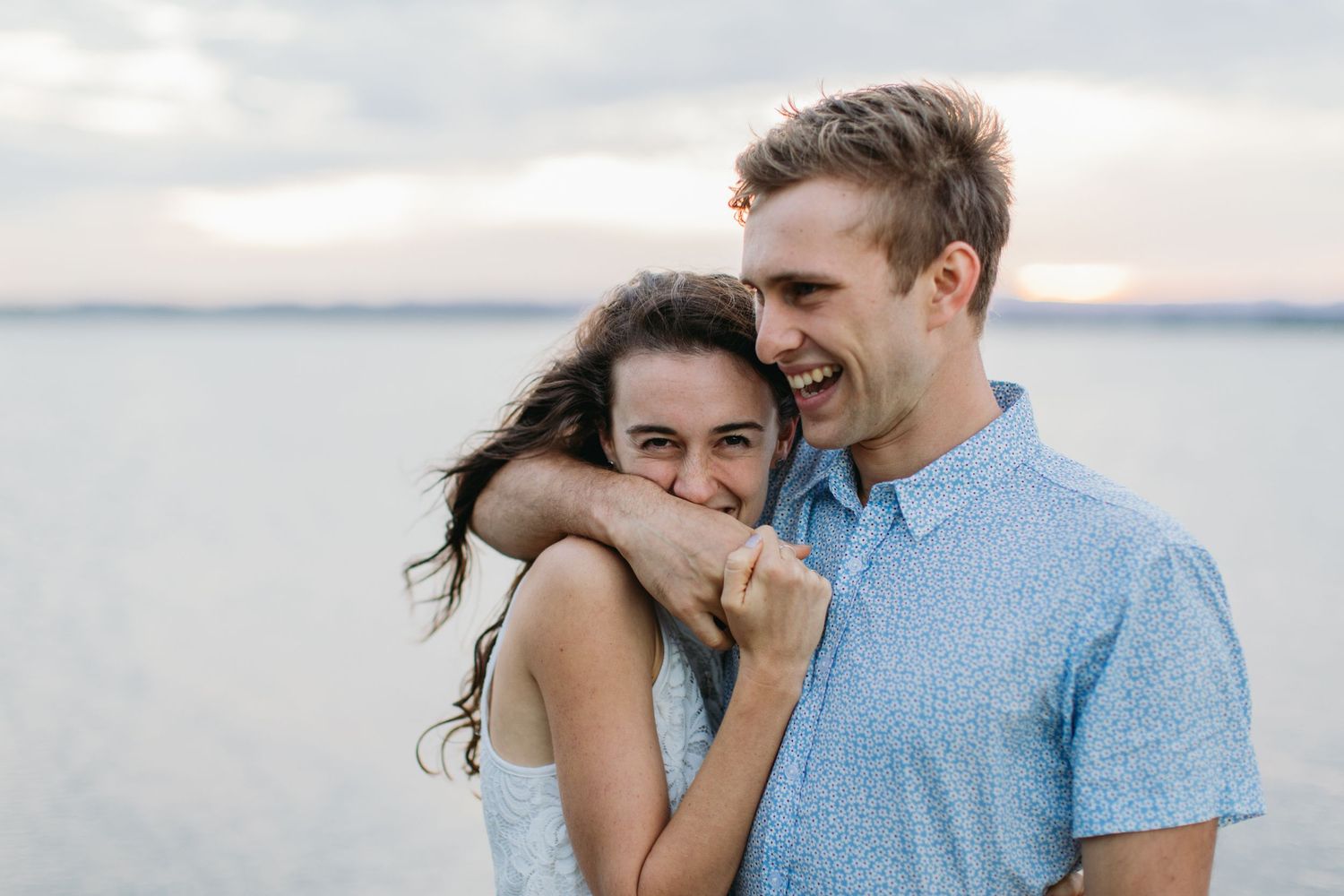Happy couple embracing and laughing together on a beach at sunset, sharing a joyful romantic moment.