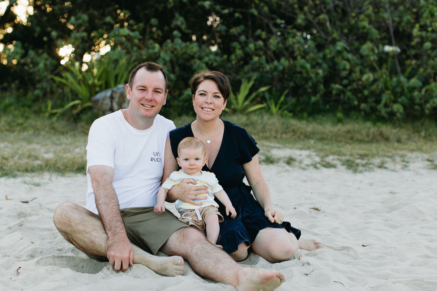 Happy family with baby sitting on sandy beach, smiling couple with infant enjoying outdoor seaside photo session.