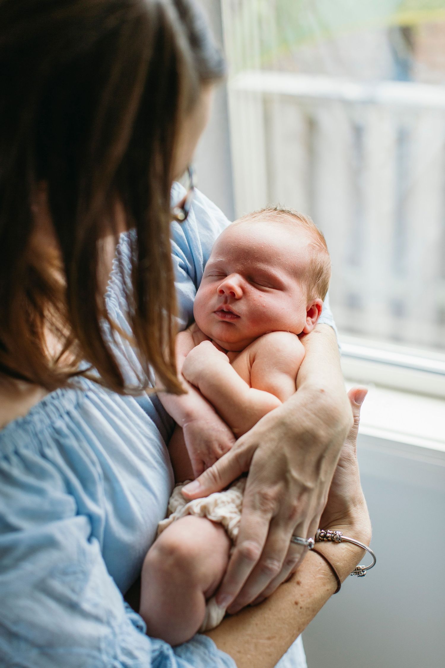 Mother in blue dress tenderly holds sleeping newborn baby wrapped in blanket near window.