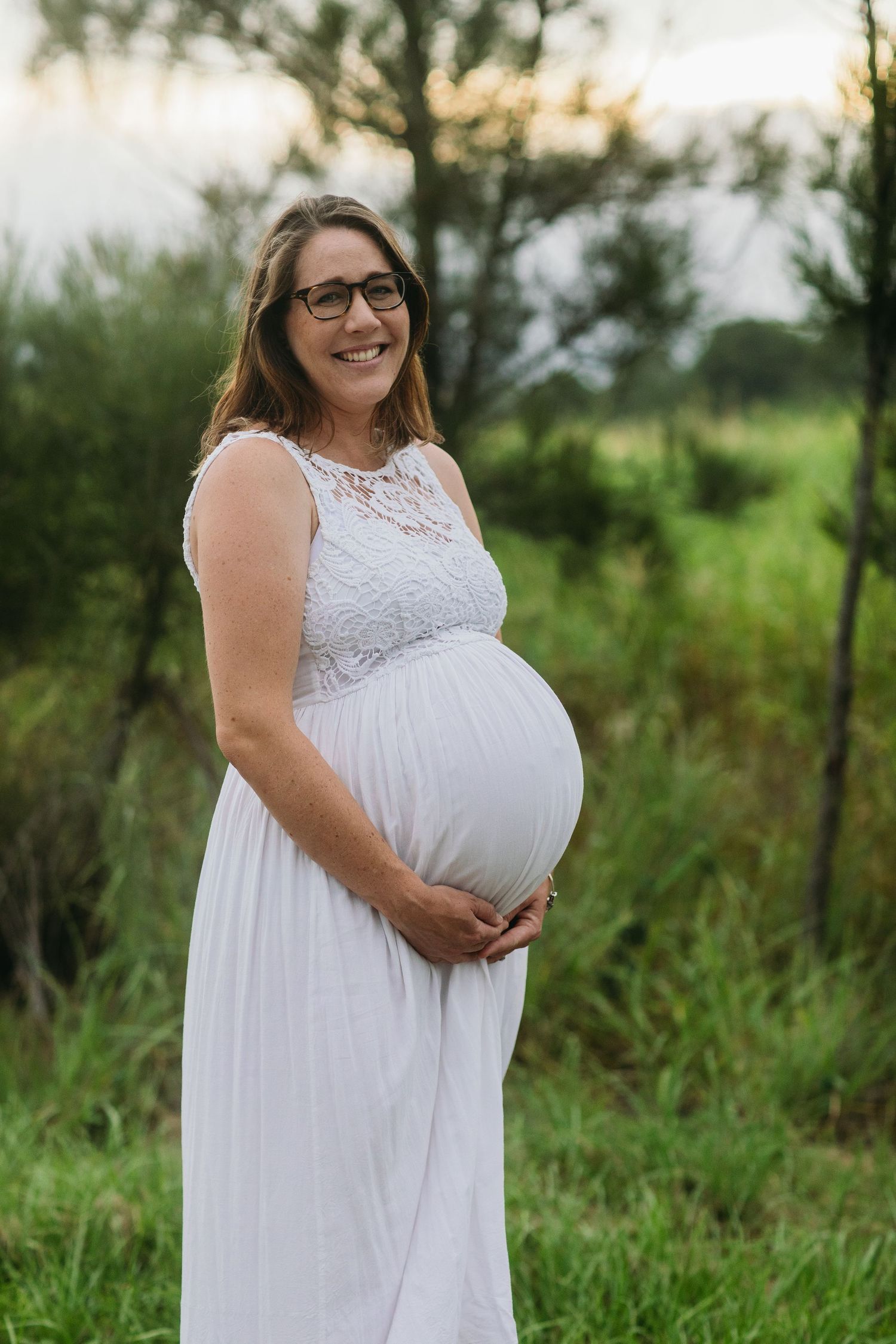 Smiling pregnant woman in white lace maxi dress holding baby bump outdoors in green nature setting.