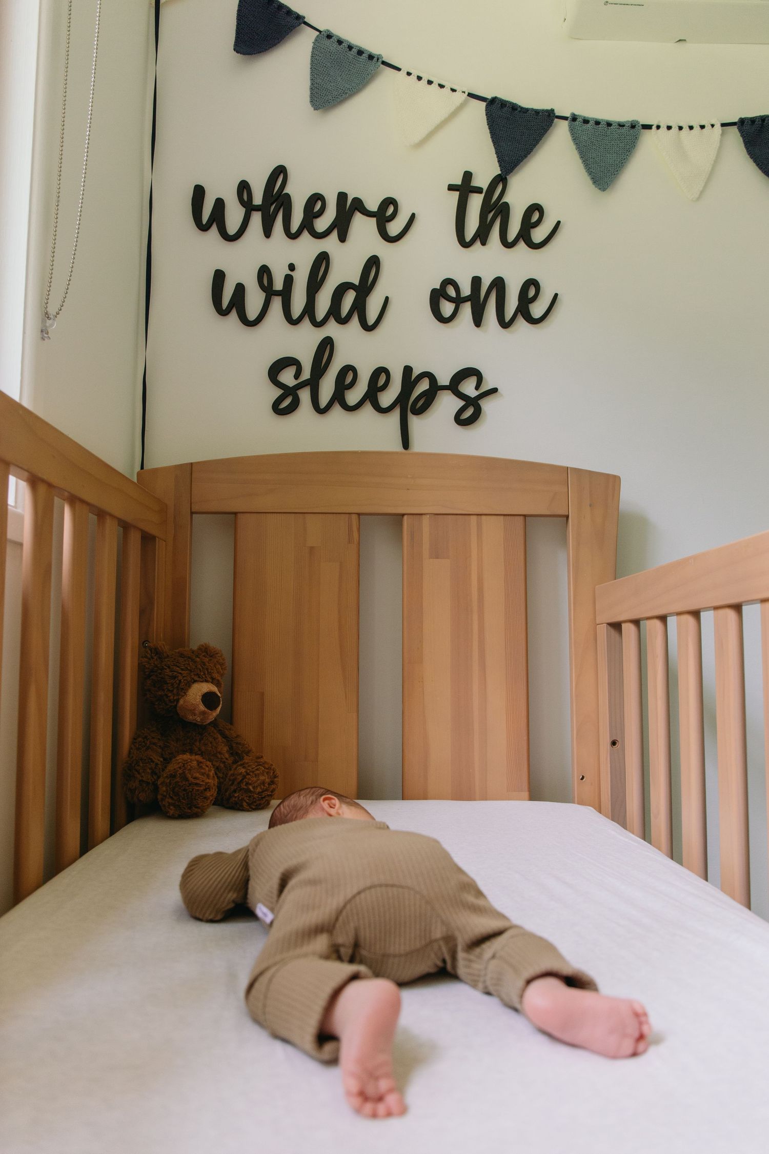 Baby sleeping in wooden crib with teddy bear, under 'where the wild one sleeps' sign and bunting.