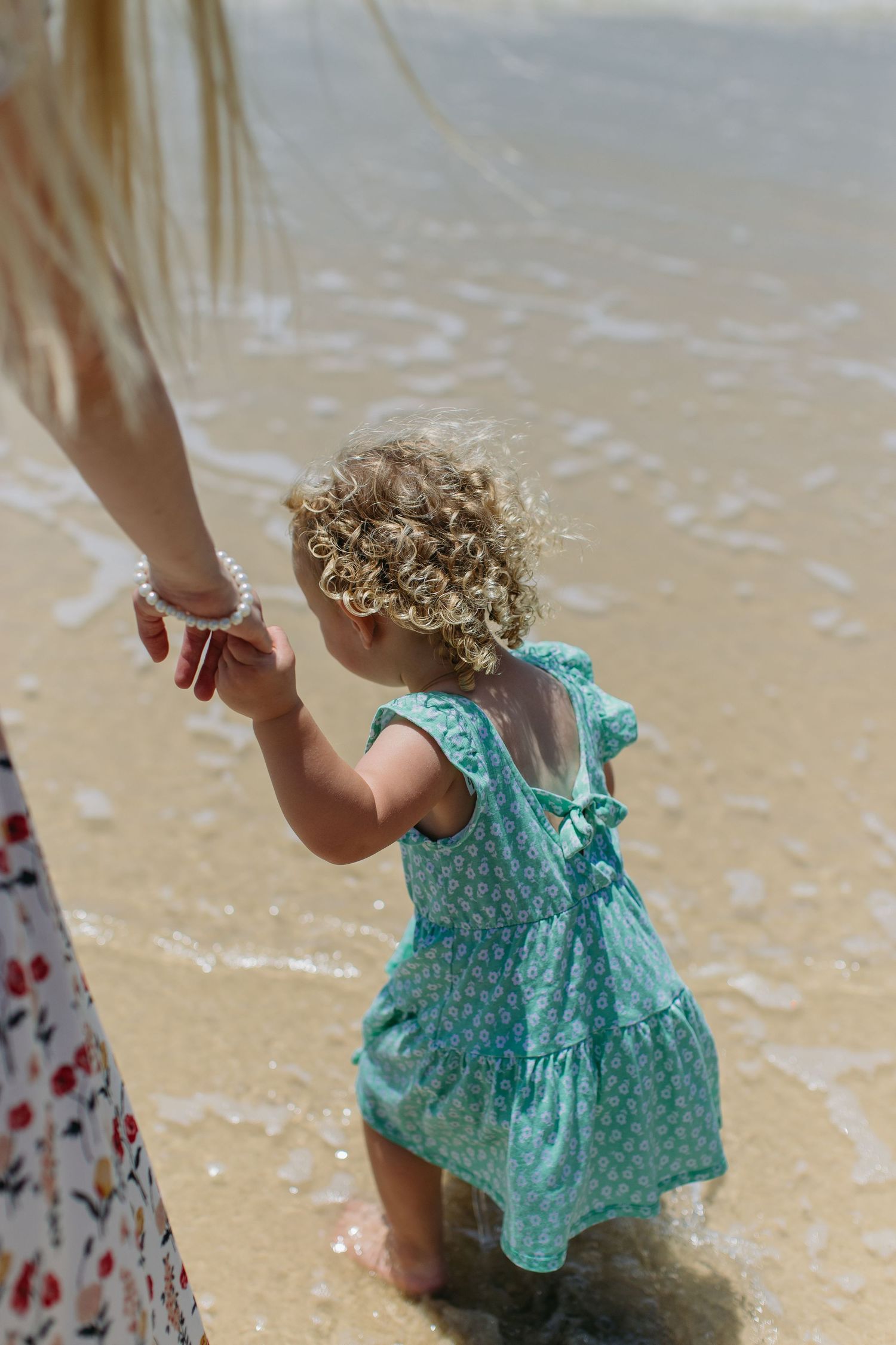 Curly-haired toddler in teal dress holding mothers hand while wading in beach shoreline.