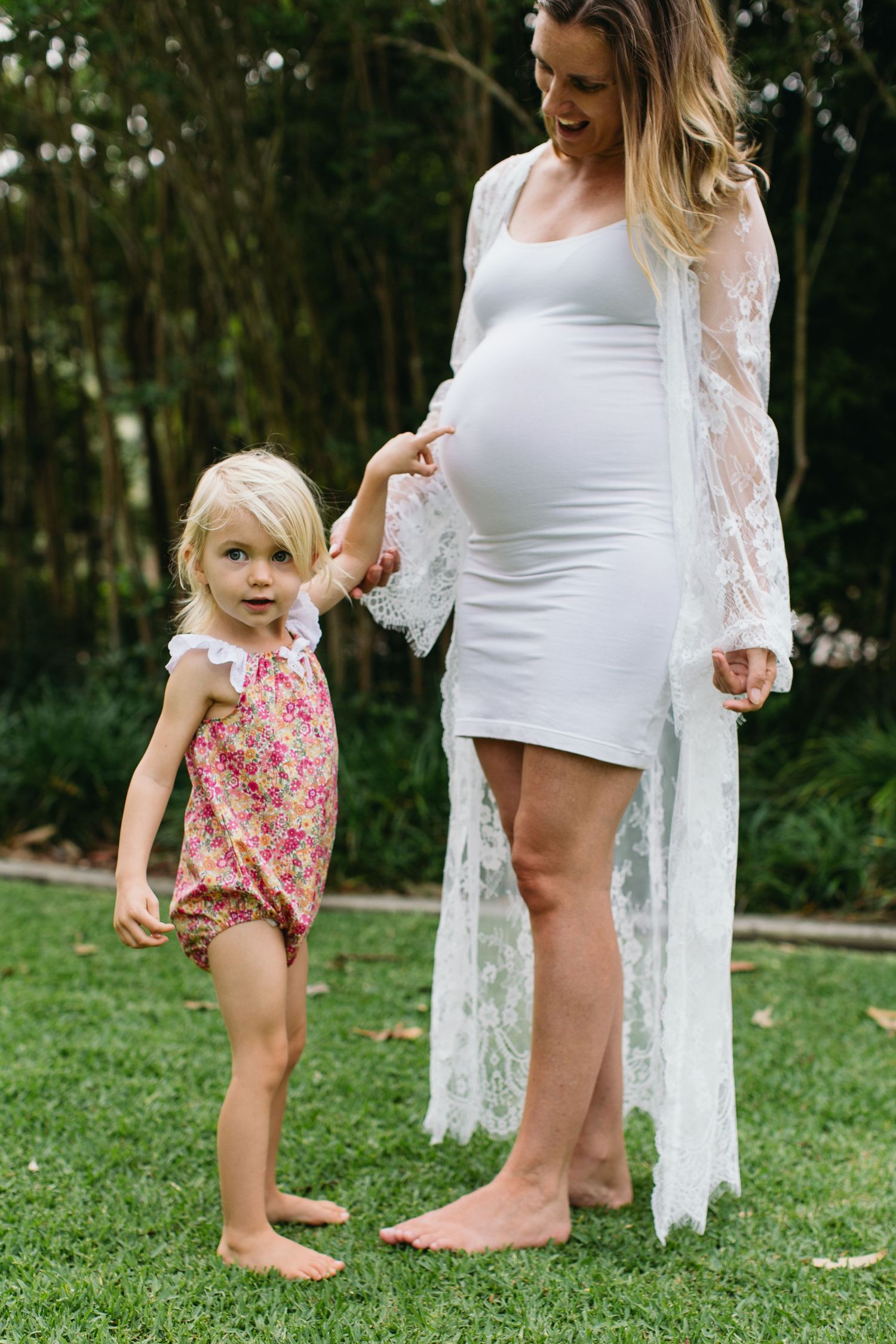Pregnant woman in white dress and lace robe stands with blonde toddler girl in floral romper on green grass.