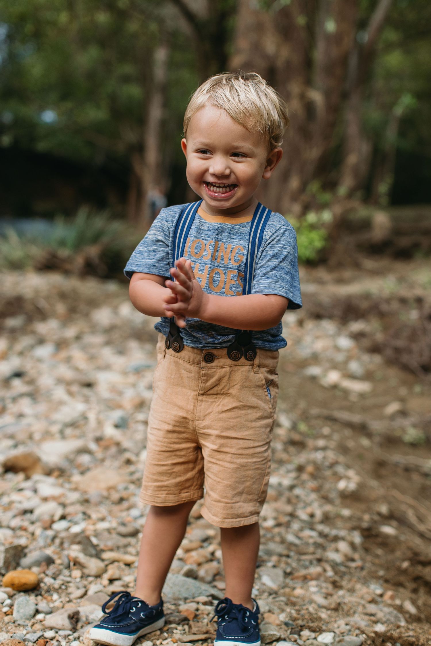 Happy toddler boy in blue shirt, suspenders, and tan shorts smiling outdoors on rocky ground.